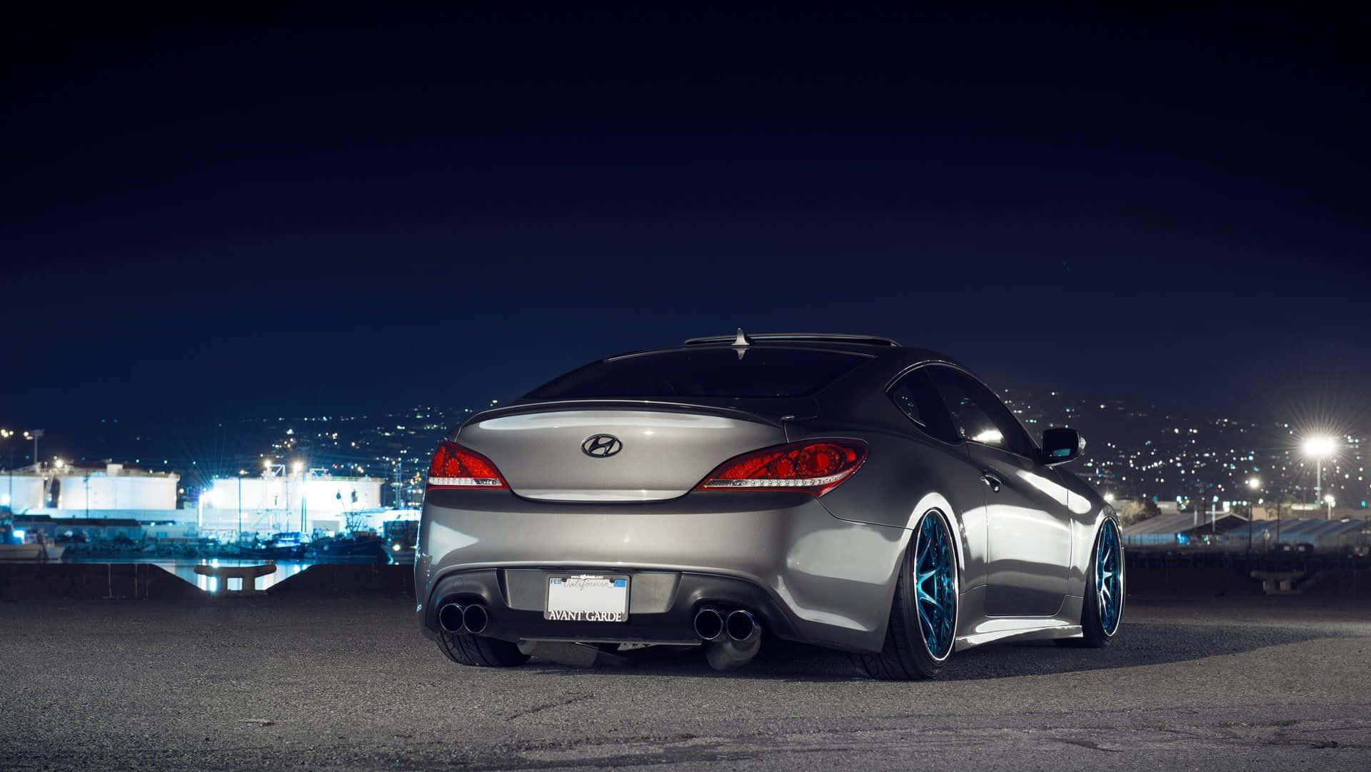 Hyundai Genesis 桌布 壁紙 — 一輛銀色現代Genesis Coupe配備定制藍色輪轂，夜晚停在天台上，俯瞰遠方燈火點亮的城市天際線。