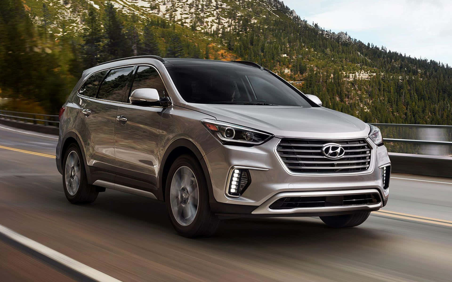 Hyundai Santa Fe 桌布 壁紙 — 一輛銀色現代SUV在高速公路飛馳，背景是森林覆蓋的山坡和積雪的山峰。