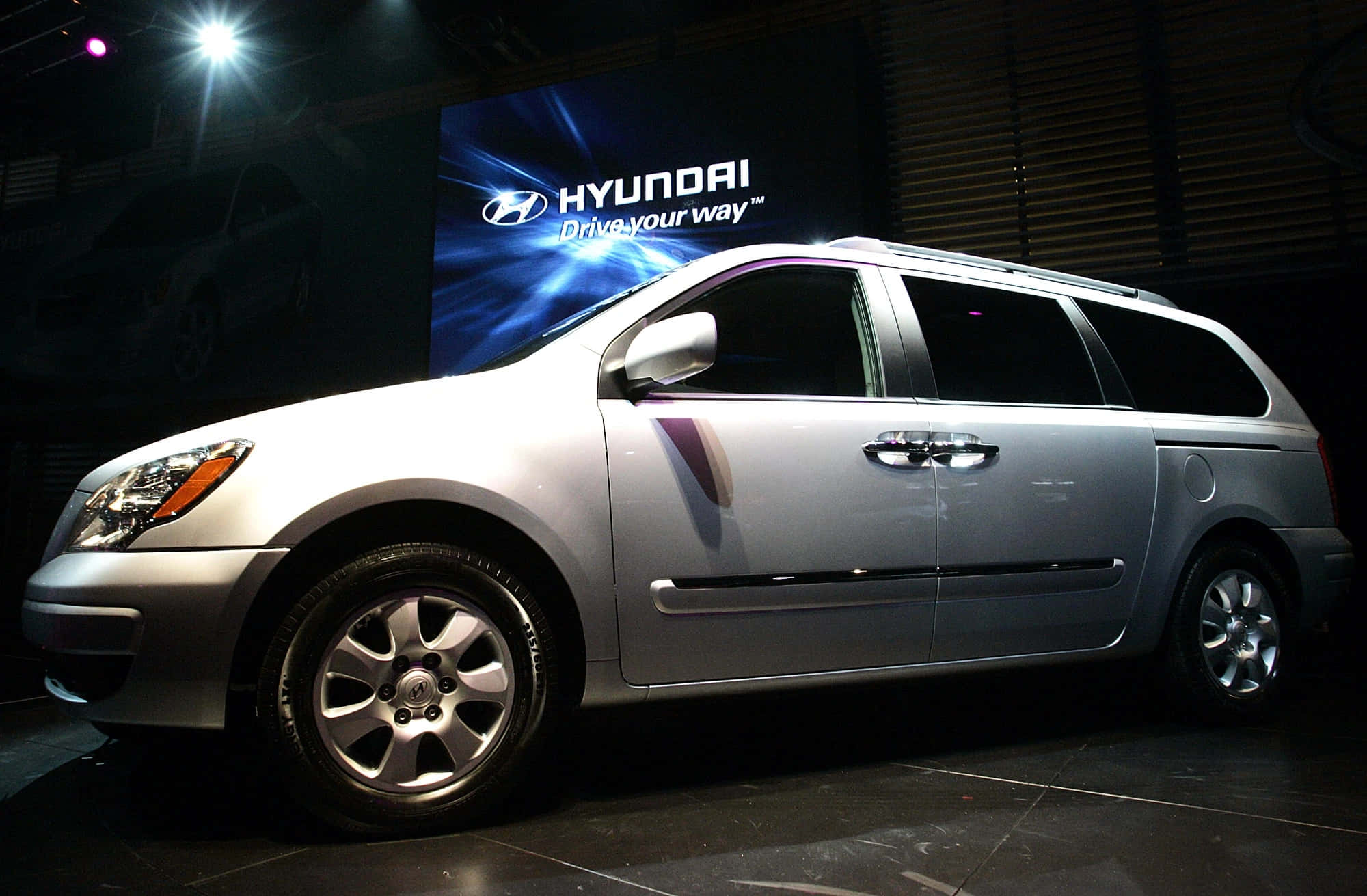 Hyundai Entourage 桌布 壁紙 — 一輛銀色現代迷你廂型車在車展的明亮舞台燈光下閃閃發光，背景帶有品牌標誌。