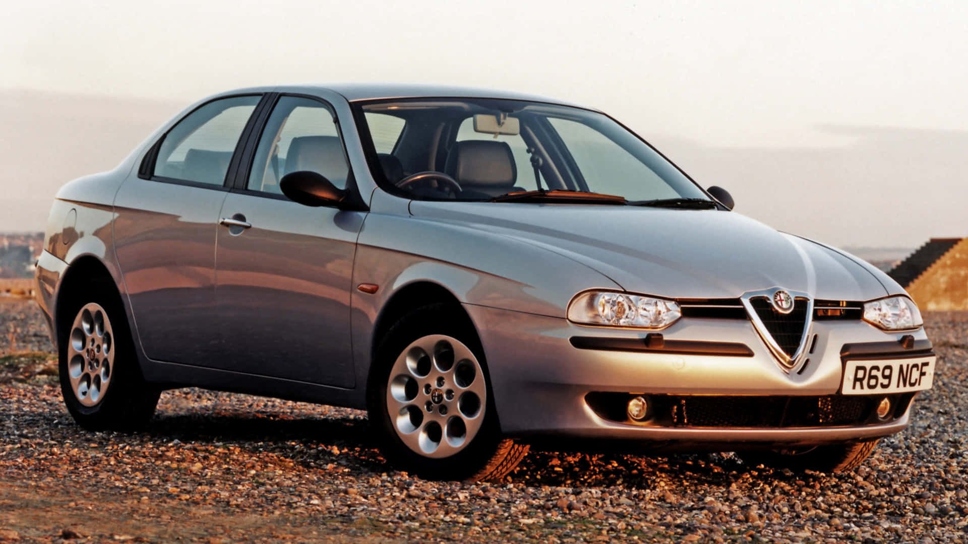 Alfa Romeo 156 桌布 壁紙 — 一輛銀色的Alfa Romeo 156轎車停在碎石場上，在柔和的黃金時段光線下。