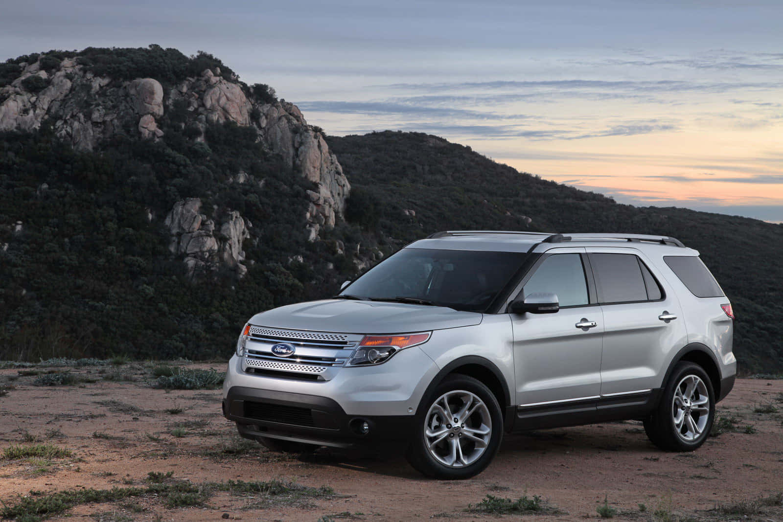 Ford Explorer 桌布 壁紙 — 一輛銀色福特 Explorer SUV 停泊在塵土飛揚的地形上，背景有岩石丘陵和柔和的日落天空。