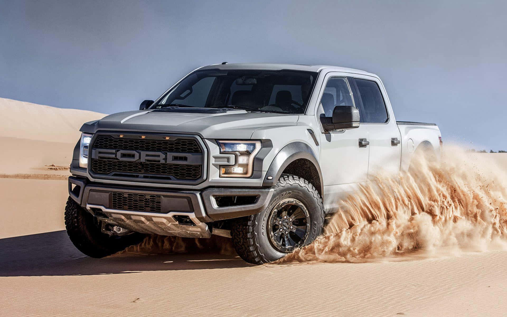 Truck 桌布 壁紙 — 一輛銀色福特 F-150 Raptor 卡車在晴朗藍天下穿越沙漠沙丘，揚起巨大沙塵。