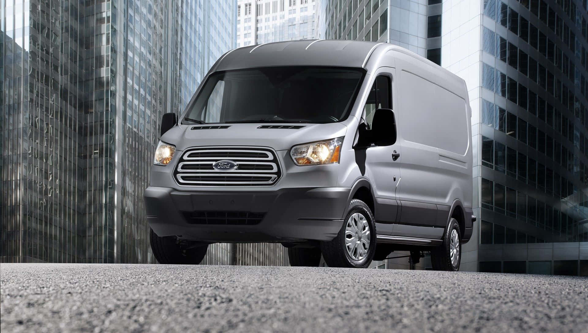 Ford Transit 桌布 壁紙 — 一輛銀色福特 Transit 客貨車停放在現代城市中高聳玻璃摩天大樓之間的柏油路面上。