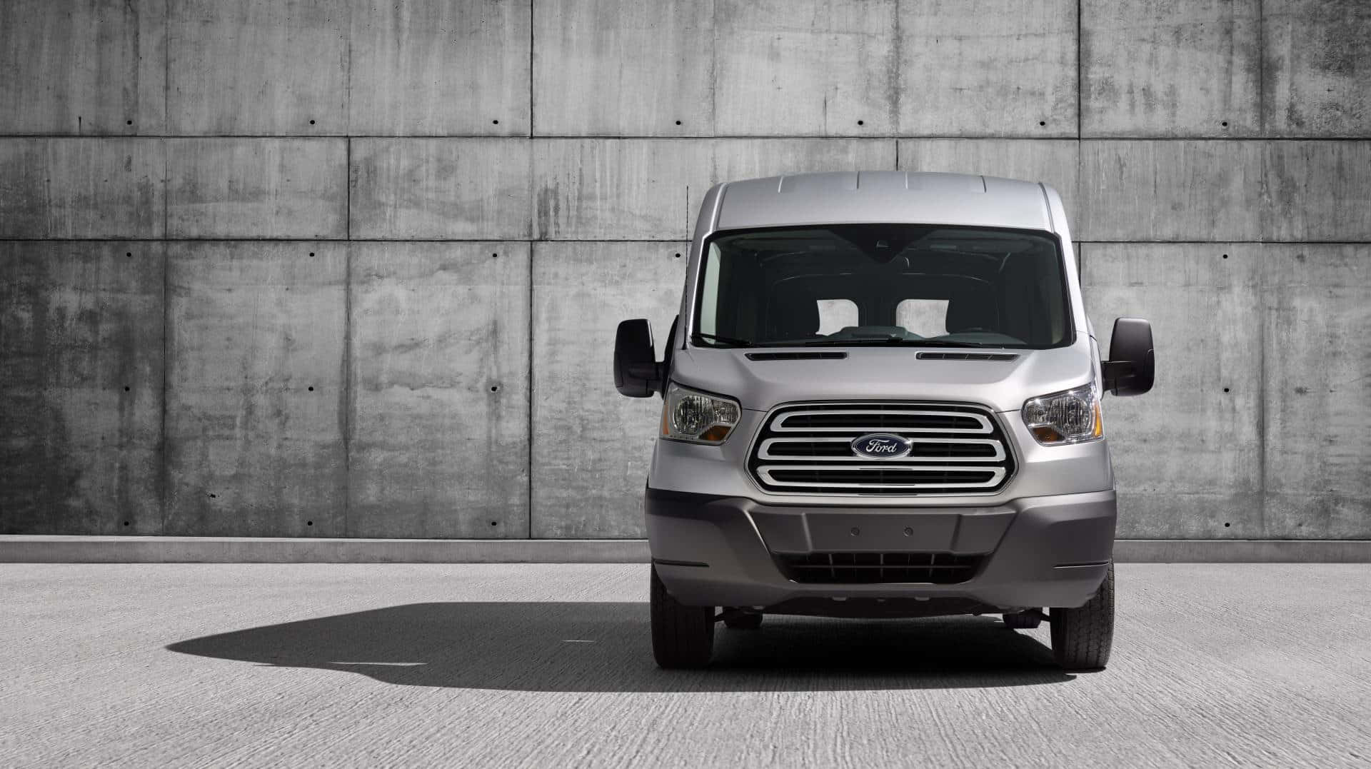 Ford Transit 桌布 壁紙 — 一輛銀色福特 Transit 廂型車在明亮的日光下，面向一面有紋理的混凝土牆。