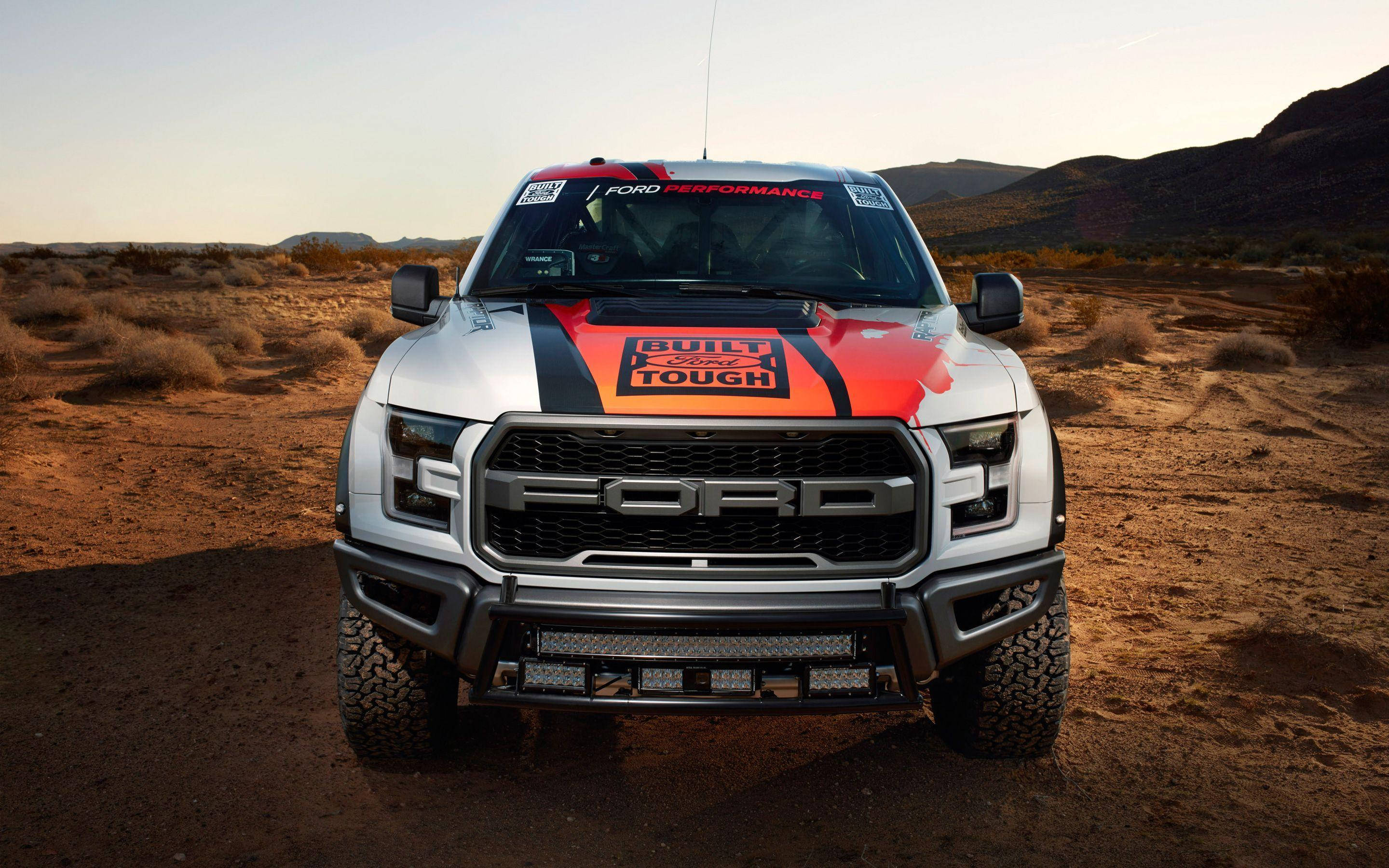 Raptor 桌布 壁紙 — 一輛銀色福特F-150 Raptor帶有橙色和黑色賽車貼花，在陽光明媚的沙漠景觀中傲然矗立。