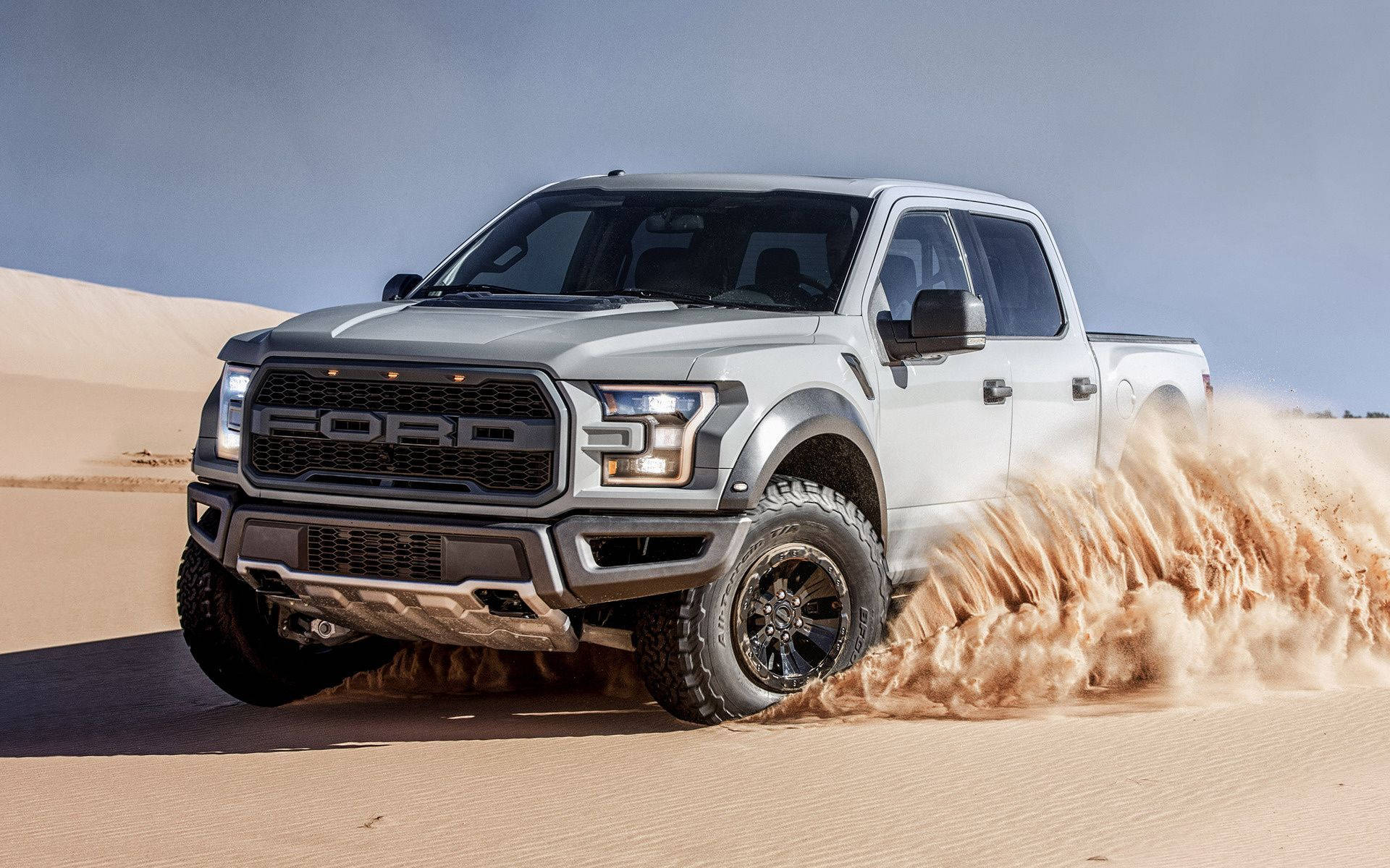 Raptor 桌布 壁紙 — 一輛銀色福特F-150猛禽卡車在晴朗的藍天下穿越沙漠沙丘，揚起巨大的沙塵雲。