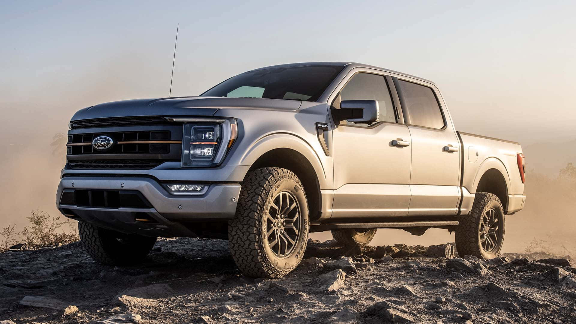 Ford F 150 桌布 壁紙 — 一輛銀色福特F-150皮卡車配備黑色輪胎，在岩石地形上行駛，揚起灰塵，天空朦朧。