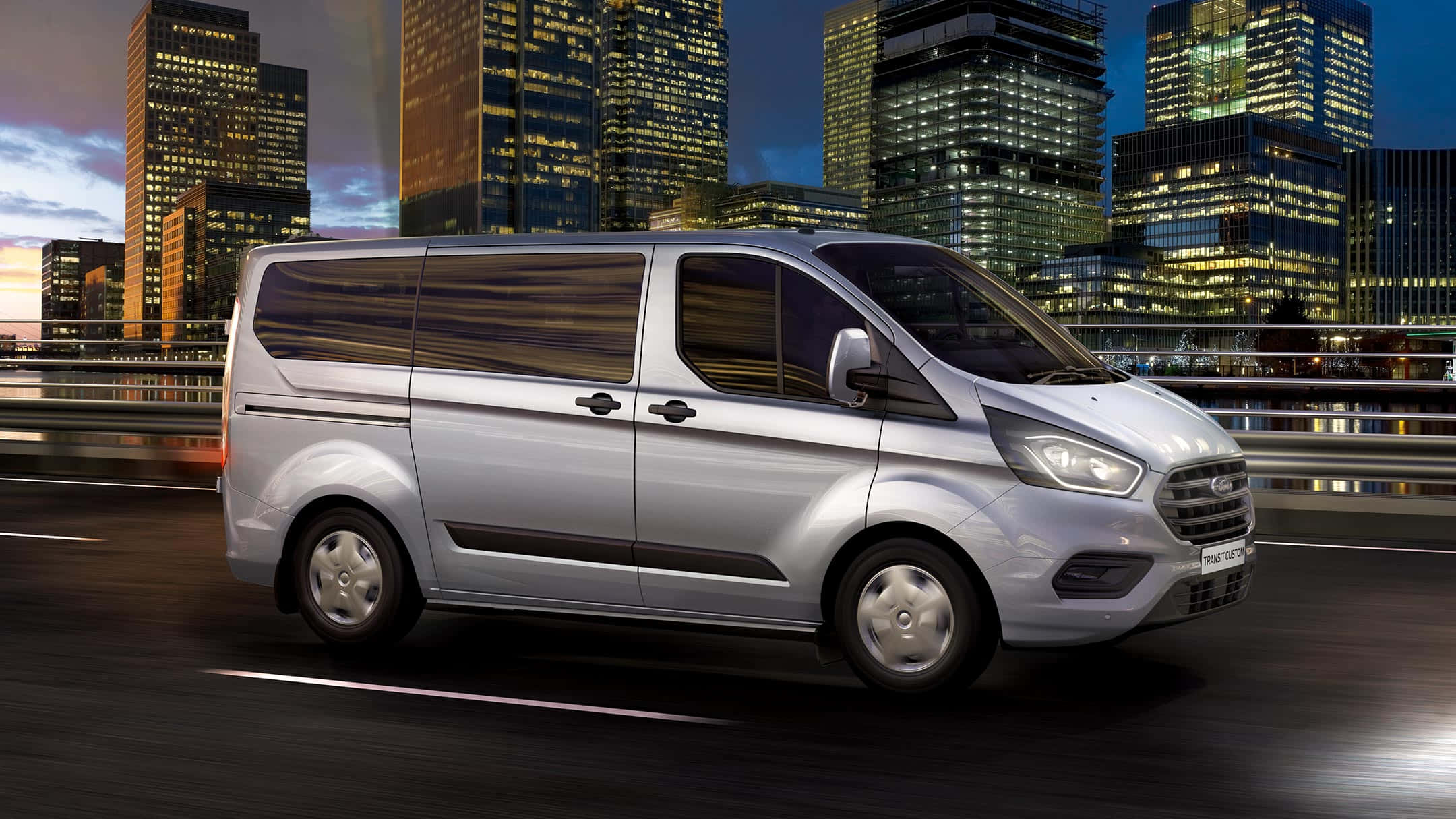 Ford Transit 桌布 壁紙 — 一輛銀色福特Transit廂型車在黃昏時分於城市高速公路上疾駛，背景是燈火通明的摩天大樓。