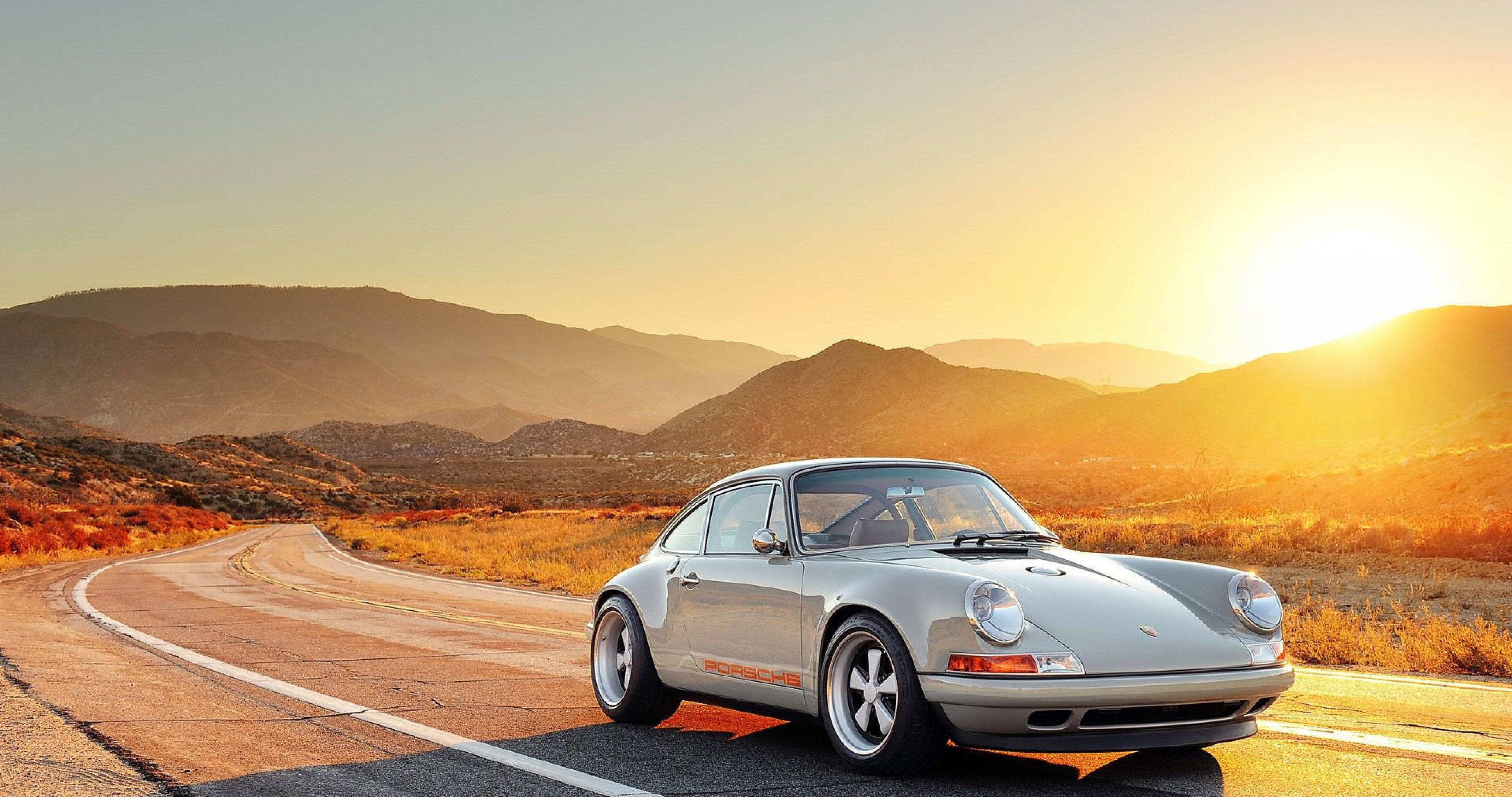 Singer 桌布 壁紙 — 一輛銀色經典保時捷911停泊在蜿蜒的沙漠公路上，背景是金色日落和山脈。