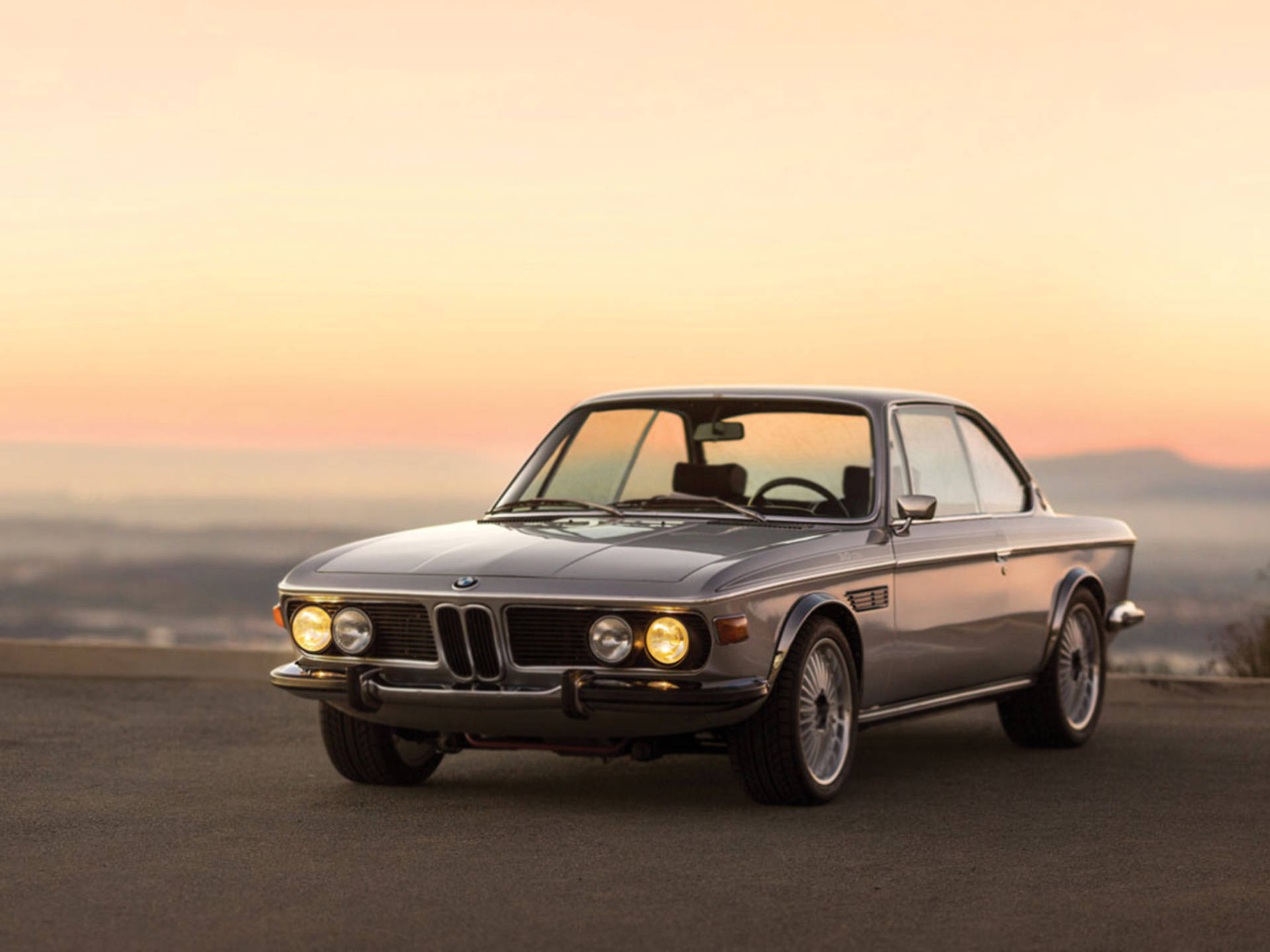 classic bmw 桌布 壁紙 — 一輛銀色經典寶馬轎跑車在溫暖的日落光線下，在背景有霧氣籠罩的山路上閃閃發光。