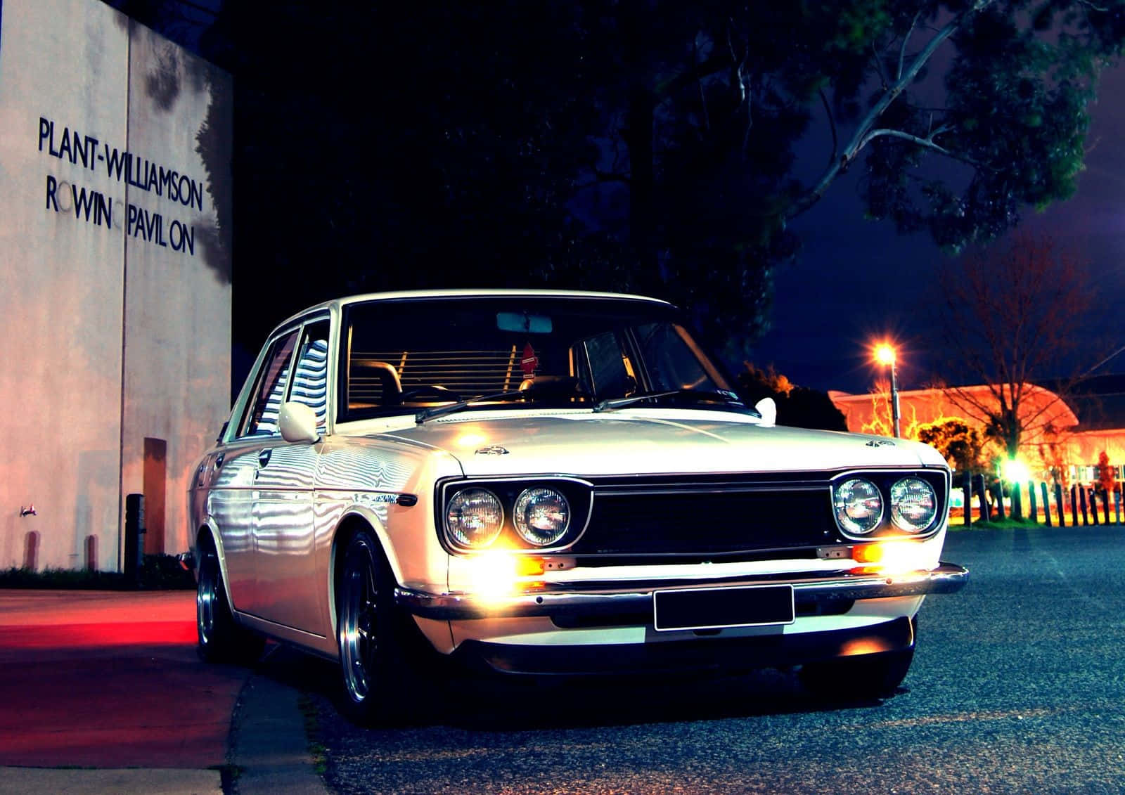 Datsun 桌布 壁紙 — 一輛銀色經典汽車在夜間停泊在設有照明標誌的建築物旁，在街燈下閃閃發光。