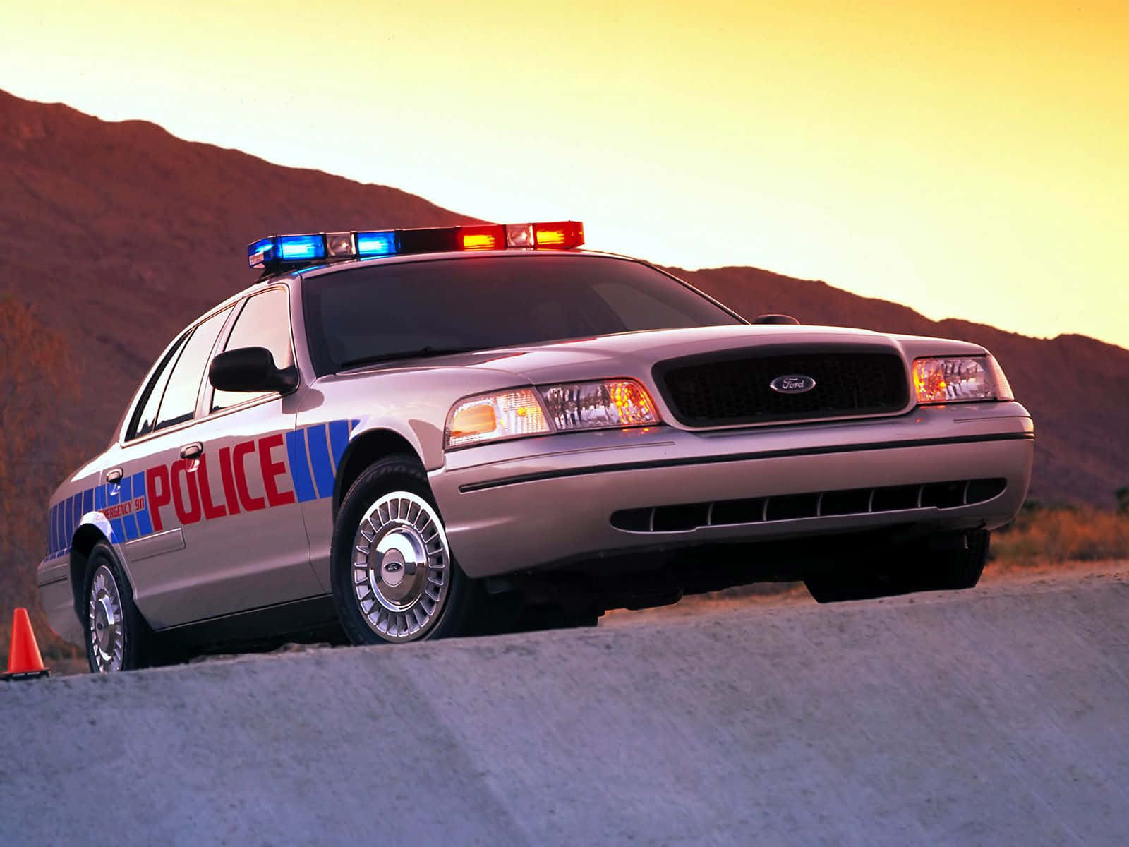 Ford Crown Victoria 桌布 壁紙 — 一輛銀色警車閃著警燈，在日落時分停靠在路邊，背景是山脈。