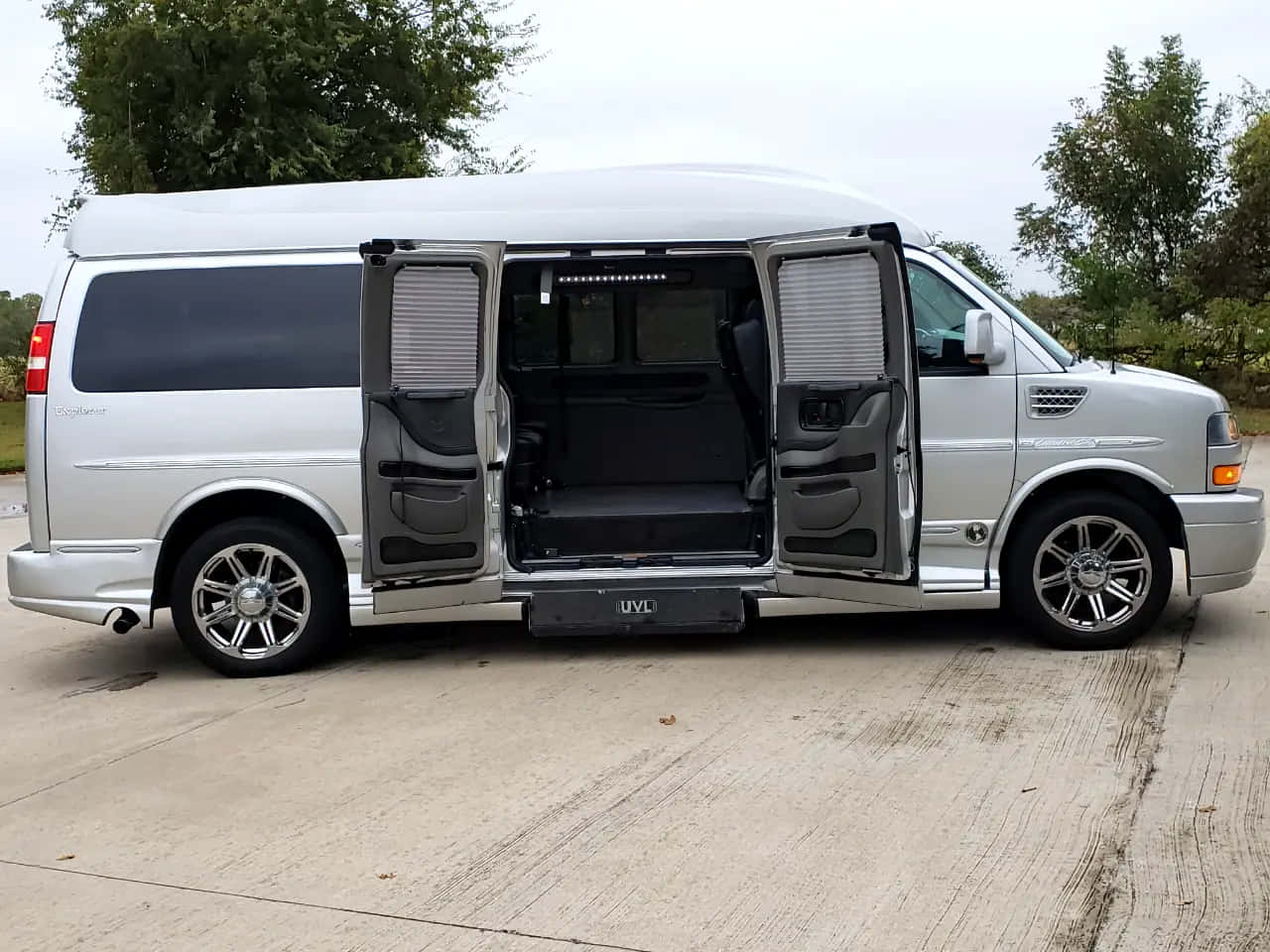 Gmc Savana 桌布 壁紙 — 一輛銀色豪華廂型車打開側門，露出黑色皮革內裝，停放在樹木環繞的混凝土車道上。