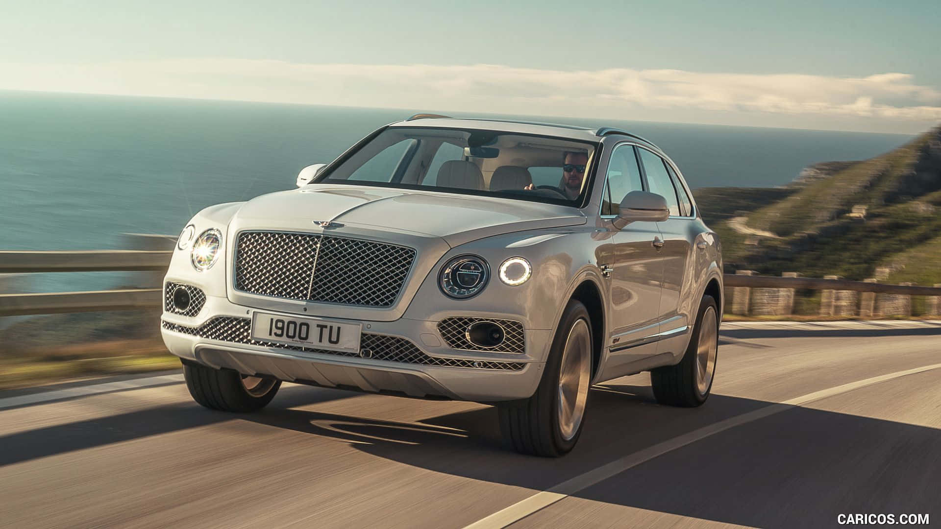 賓利 桌布 壁紙 — 一輛銀色賓利 Bentayga SUV 沿著海岸公路行駛，背景是海洋和懸崖，天空晴朗。
