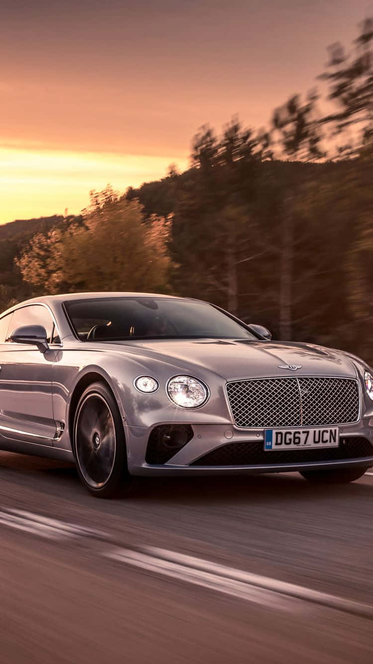 bentley HD 桌布 壁紙 — 一輛銀色賓利 Continental GT 在日落時分沿著道路疾馳，背景是模糊的樹木和溫暖的金色光線。