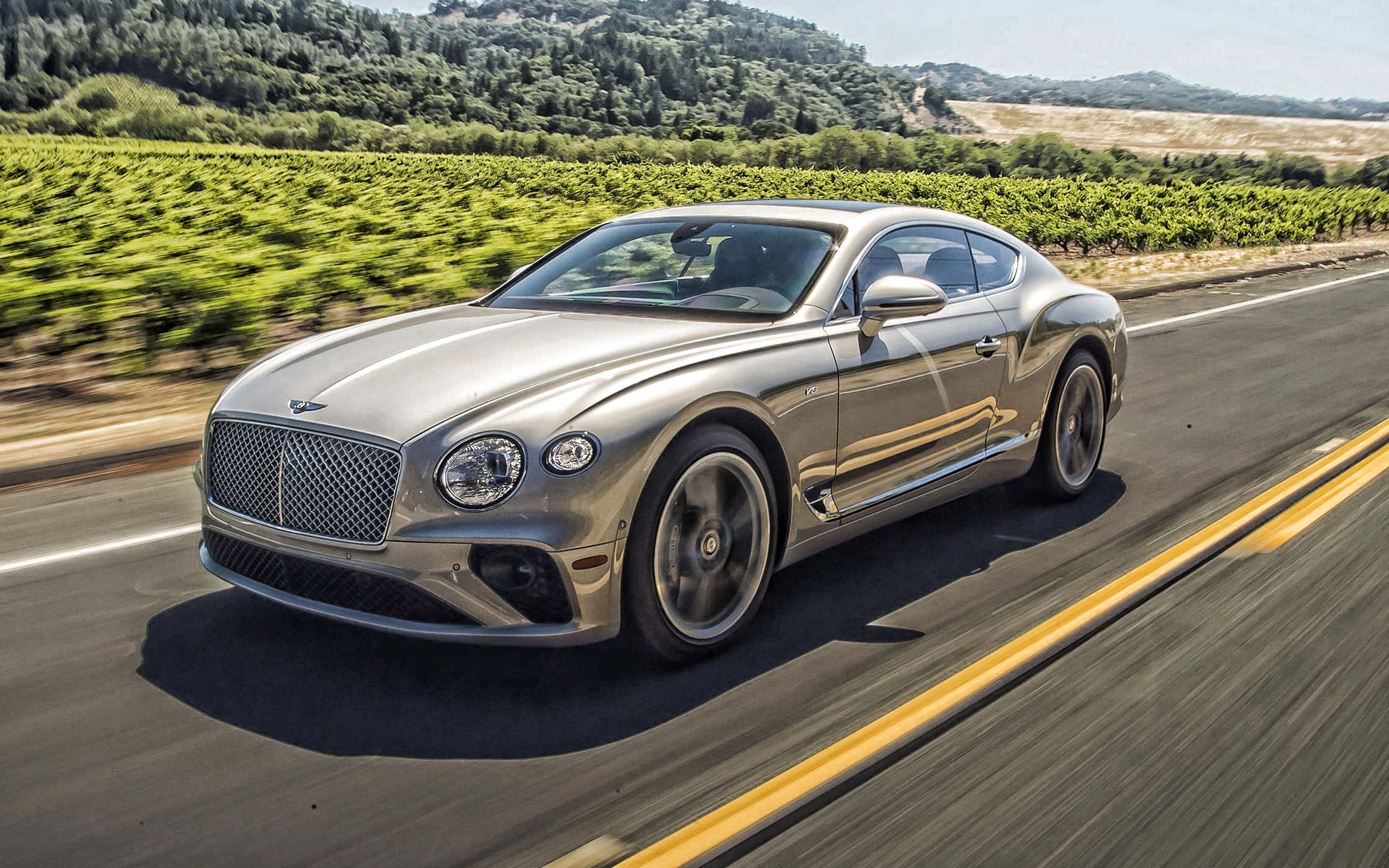 桌布 壁紙 — 一輛銀色賓利 Continental GT 在陽光明媚的道路上行駛，兩旁是鬱鬱蔥蔥的綠色葡萄園和起伏的丘陵。