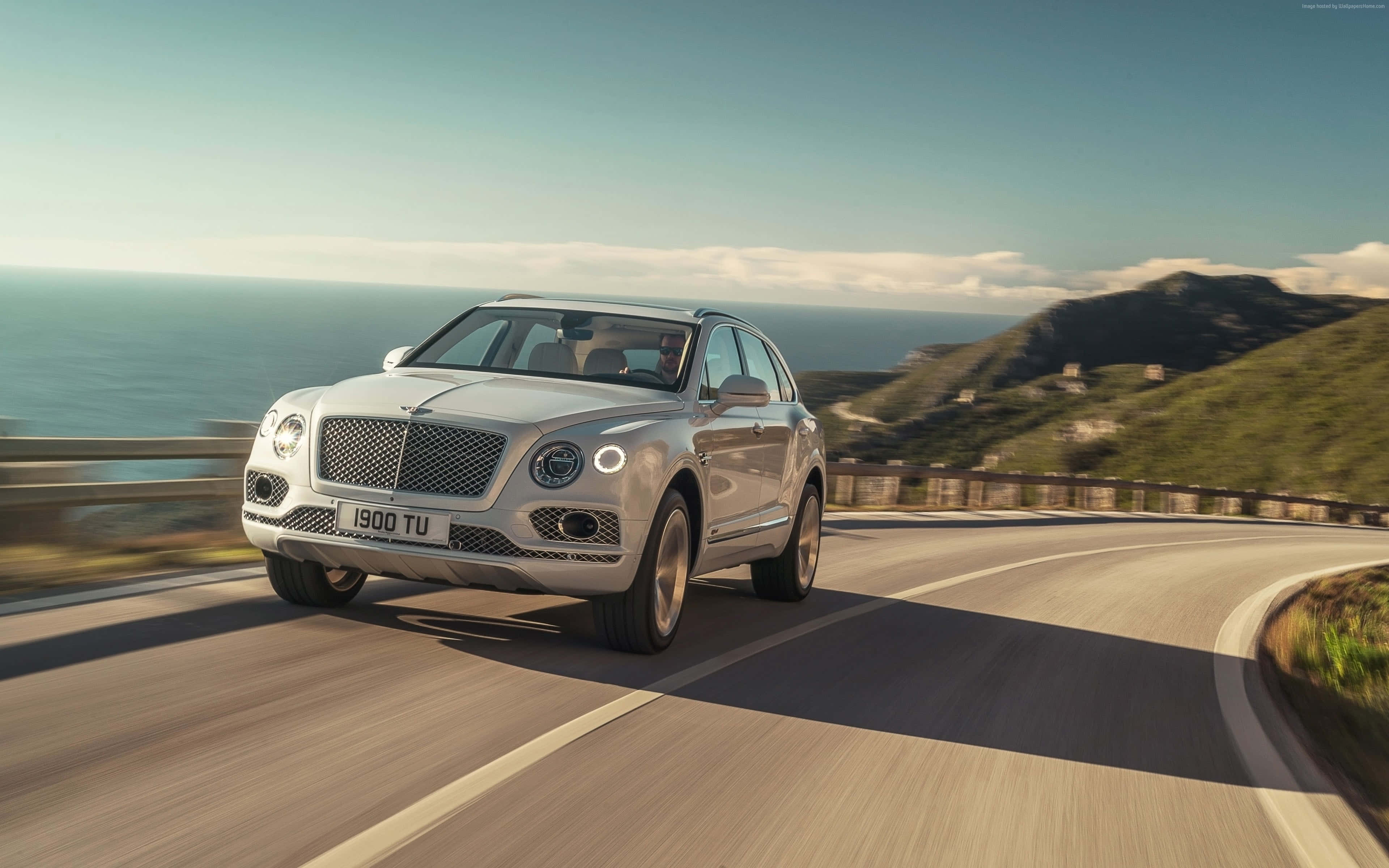 Bentley Bentayga 桌布 壁紙 — 一輛銀色賓利Bentayga SUV沿著海岸山路行駛，背景是海洋和綠色山丘，天空晴朗。