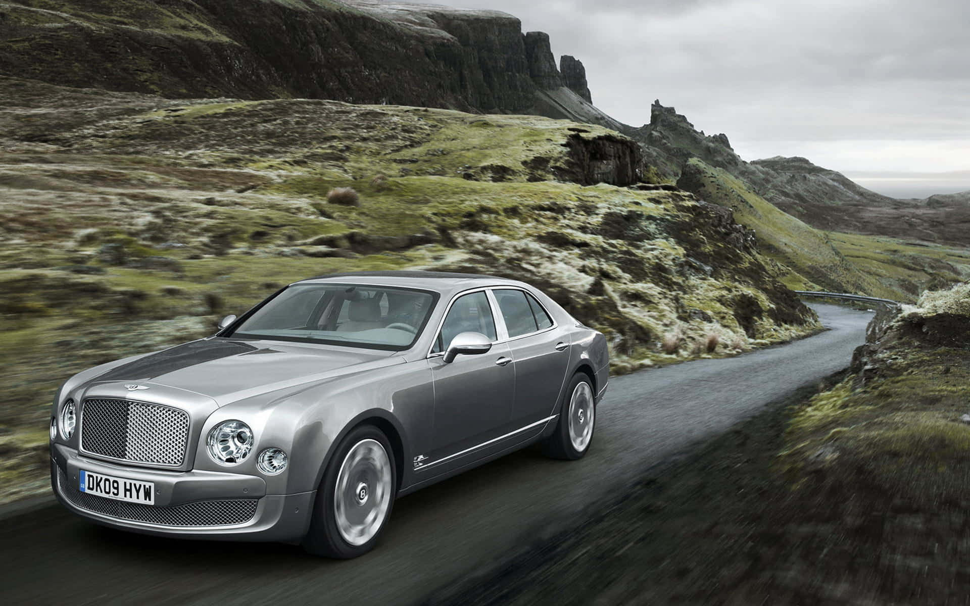 Bentley Mulsanne 桌布 壁紙 — 一輛銀色賓利房車在陰天之下，沿著蜿蜒的山路穿過霧氣瀰漫的綠色山丘飛馳。