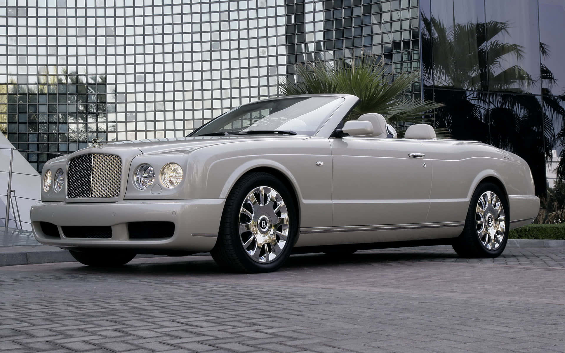Bentley Azure 桌布 壁紙 — 一輛銀色賓利敞篷車停放在鋪砌的廣場上，前方是一座玻璃摩天大樓，棕櫚樹倒映在窗戶中。