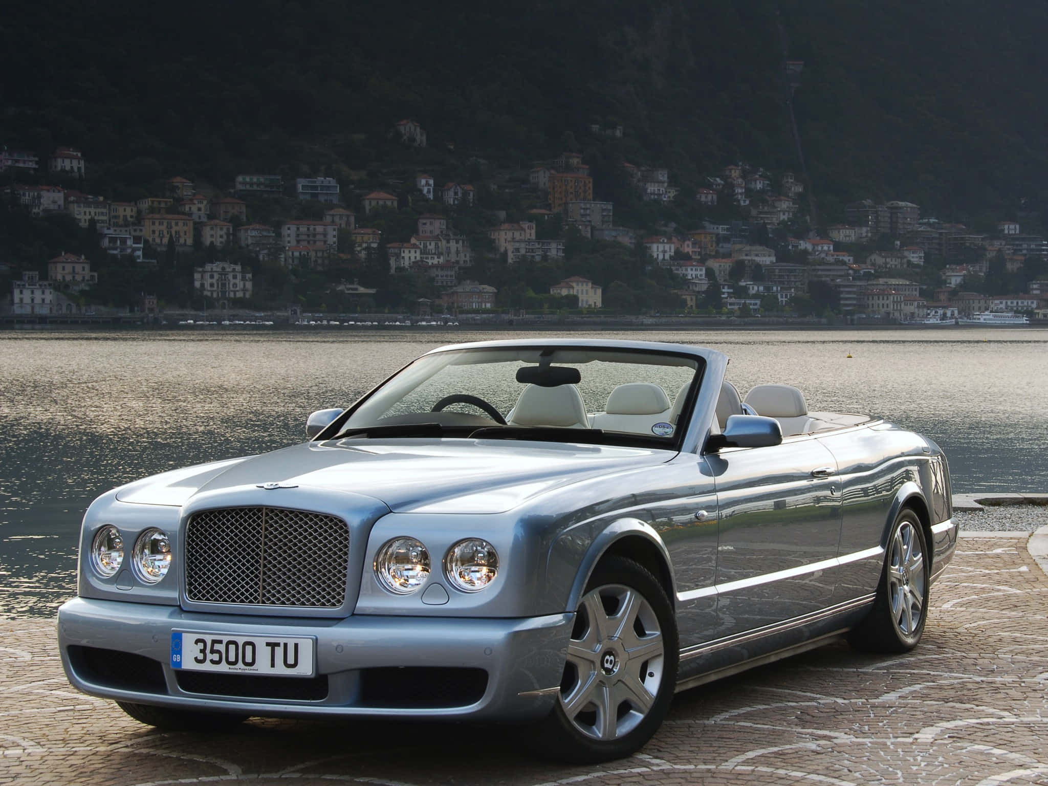 Bentley Azure 桌布 壁紙 — 一輛銀色賓利敞篷車停泊在平靜湖泊旁的石碼頭上，背景是山坡村莊。