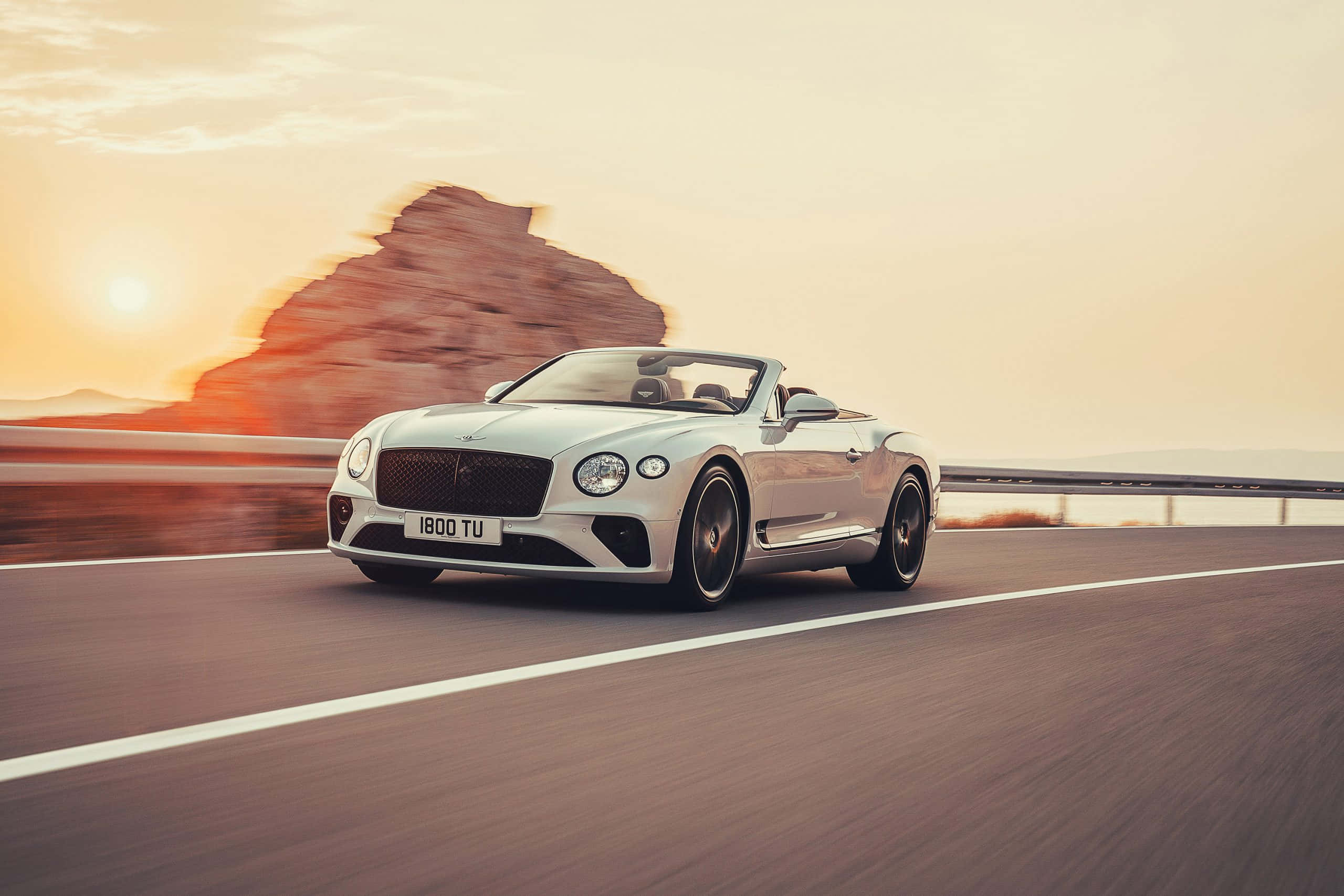 Bentley Continental Gt Convertible 桌布 壁紙 — 一輛銀色賓利敞篷車在日落時分沿沙漠高速公路疾駛，背景有模糊的岩石形成。