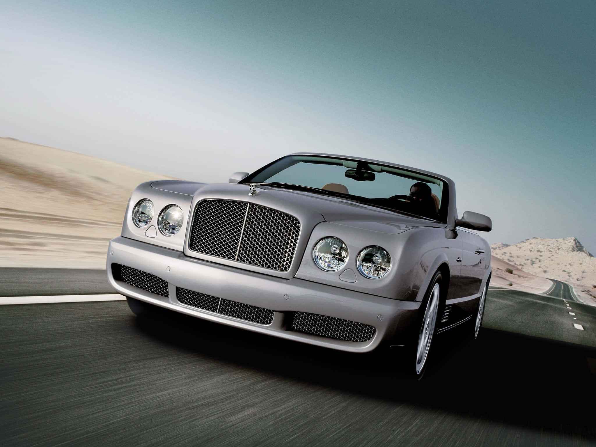 Bentley Azure 桌布 壁紙 — 一輛銀色賓利敞篷車在晴朗天空下，沿沙漠公路高速行駛，呈現模糊動感及岩石地形。