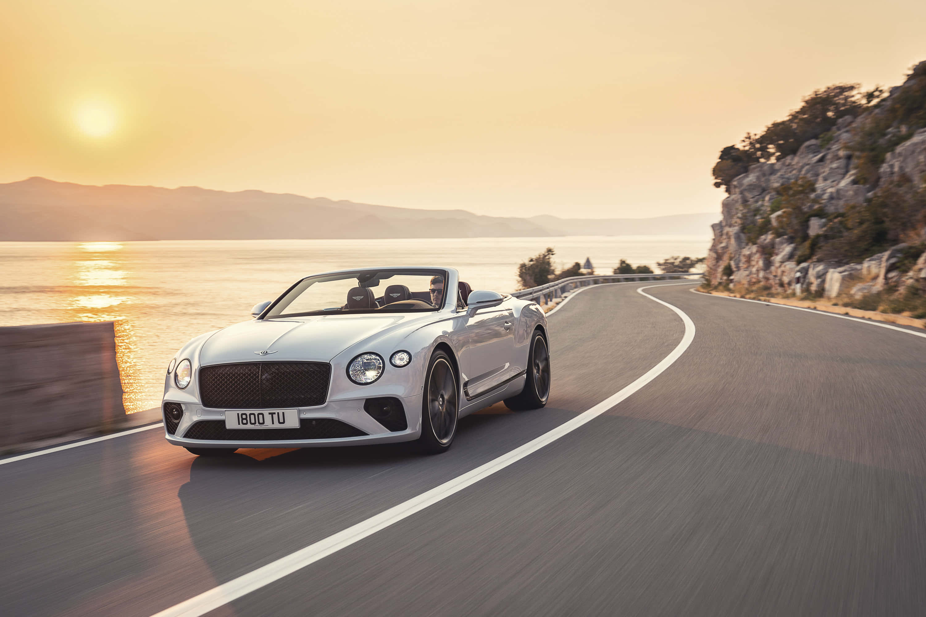 Bentley Continental Gt Convertible 桌布 壁紙 — 一輛銀色賓利敞篷車沿著海岸公路在日落時分巡航，金色光芒在水面上反射。