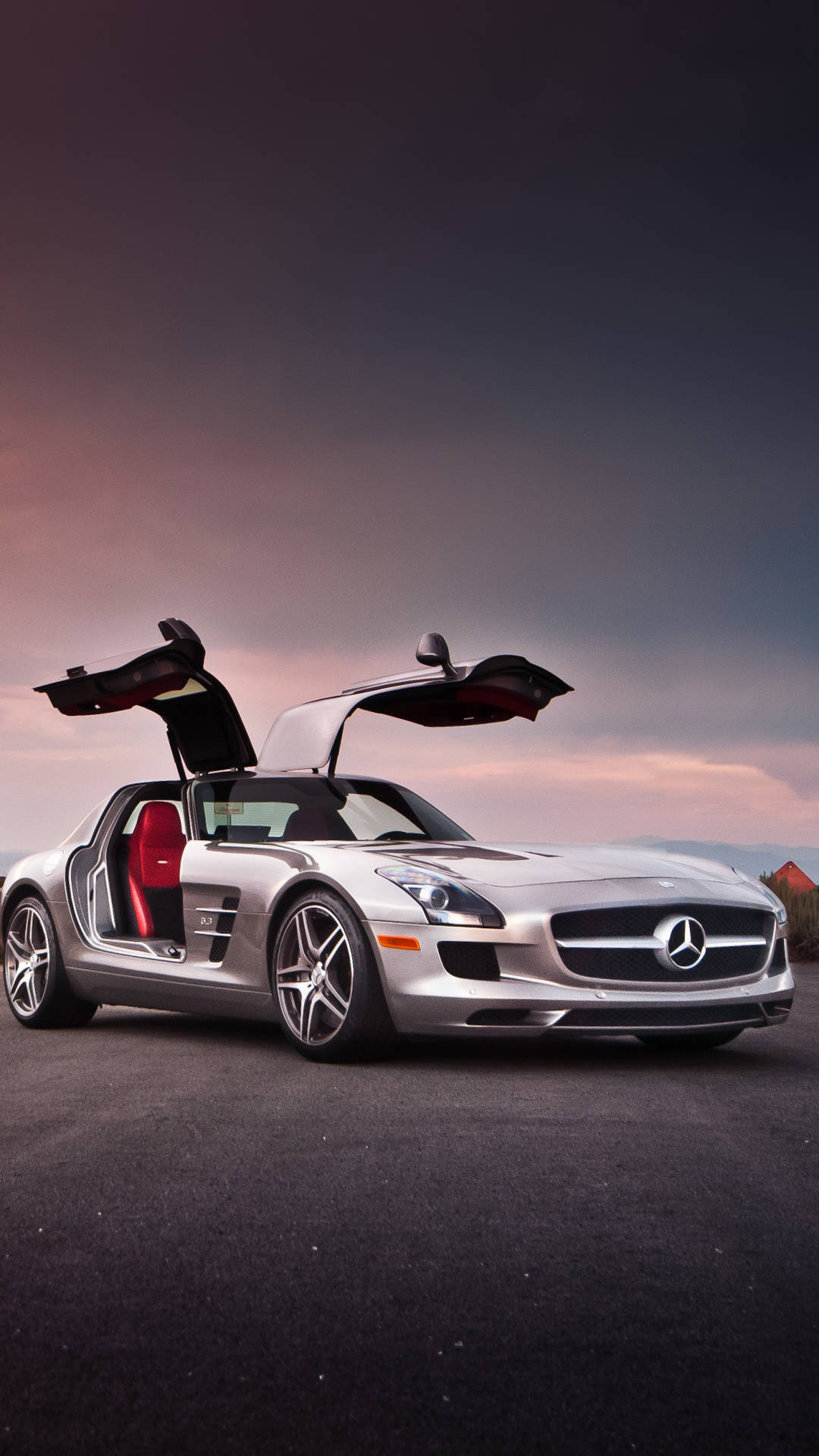 賓士 iPhone 桌布 壁紙 — 一輛銀色賓士 SLS AMG 跑車，鷗翼車門打開，停在柏油路上，背景是帶有粉紅和紫色雲彩的暮色天空。