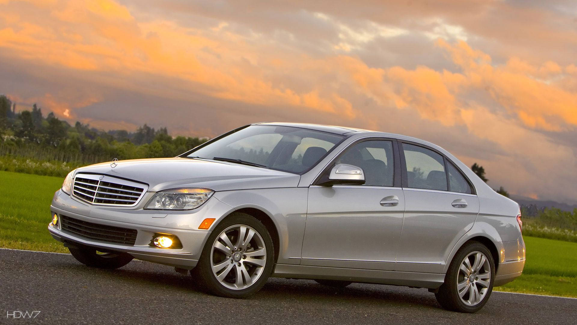 Mercedes Benz C300 桌布 壁紙 — 一輛銀色賓士轎車在金色夕陽天空下閃閃發光，天空有橙色和粉紅色的雲彩，下方是綠色田野。