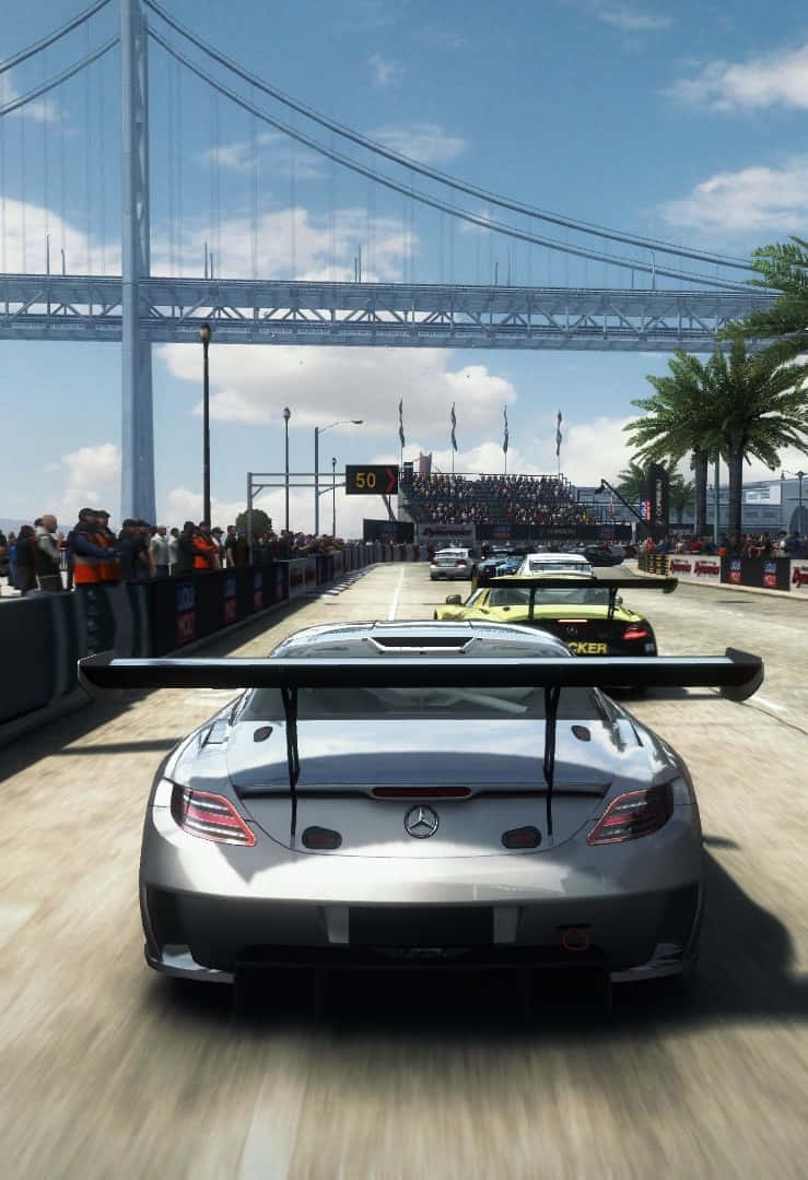iPhone X Grid Autosport 桌布 壁紙 — 一輛銀色賽車在巨型吊橋下的賽道上飛馳，觀眾在旁邊歡呼。