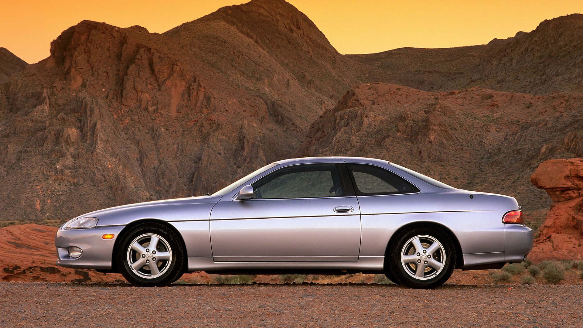 Lexus Sc 300 桌布 壁紙 — 一輛銀色跑車停泊在沙漠公路上，周圍是崎嶇的紅色岩層，上方是溫暖的橙色日落天空。