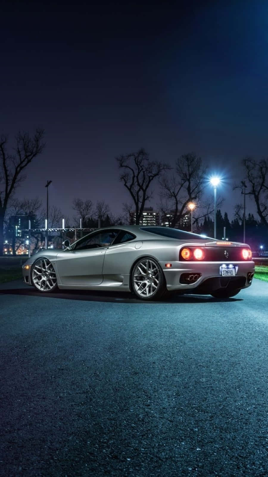 Ferrari 360 Modena 桌布 壁紙 — 一輛銀色跑車在夜晚街燈下閃耀，停泊在瀝青停車場，周圍有光禿的樹木和遠處的城市燈光。