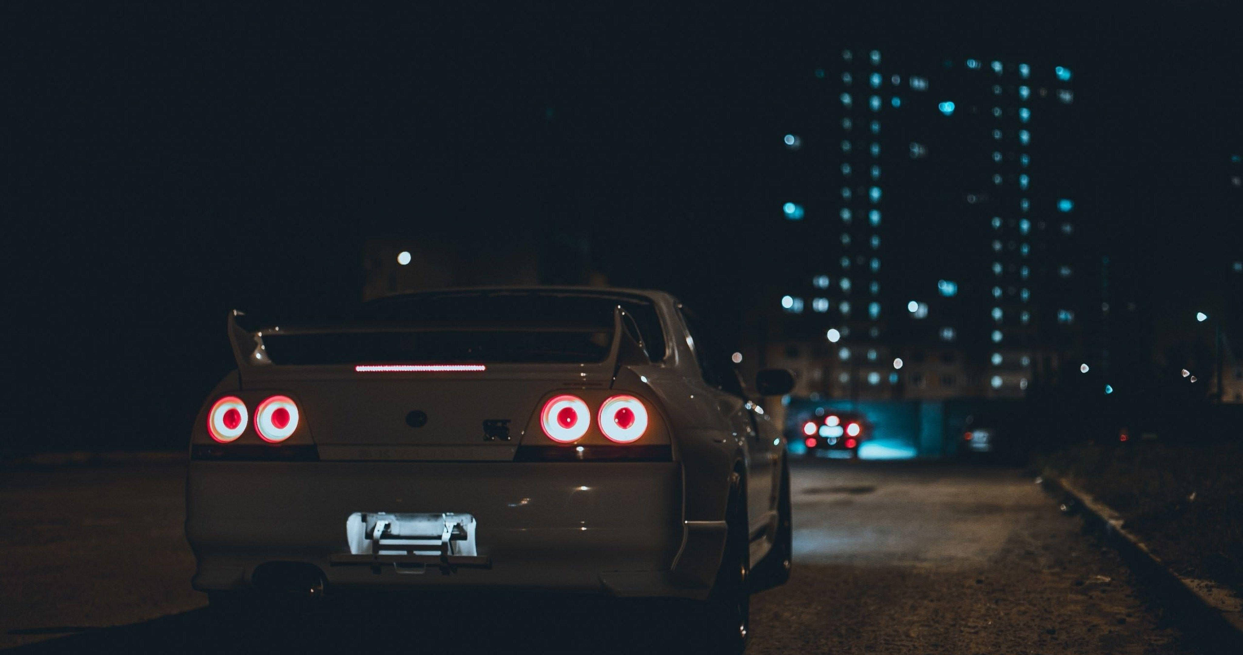 Gtr 桌布 壁紙 — 一輛銀色跑車在夜間停泊在黑暗的城市街道上，紅色尾燈閃爍，背景是燈火通明的摩天大樓。
