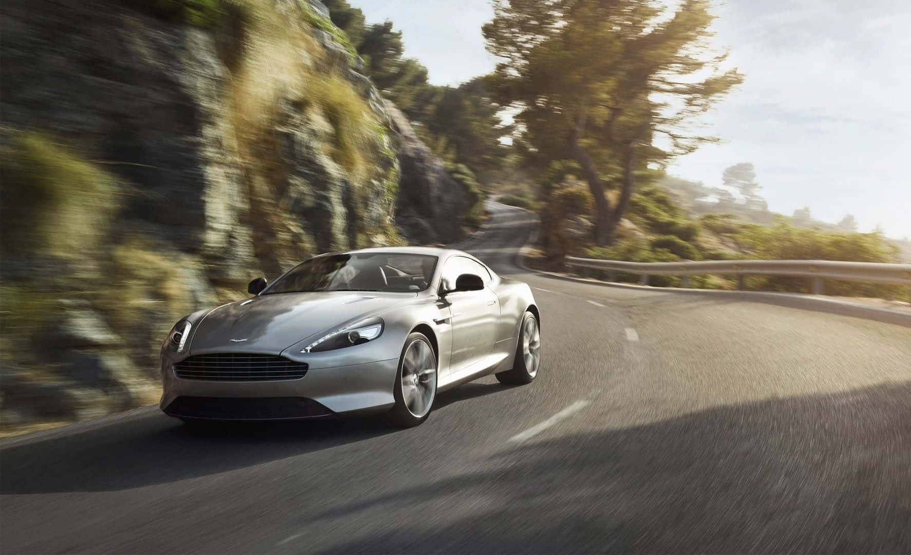 Aston Martin Db9 桌布 壁紙 — 一輛銀色跑車在明媚陽光下，沿著蜿蜒的山路飛馳，兩旁是模糊的綠樹和岩石峭壁。