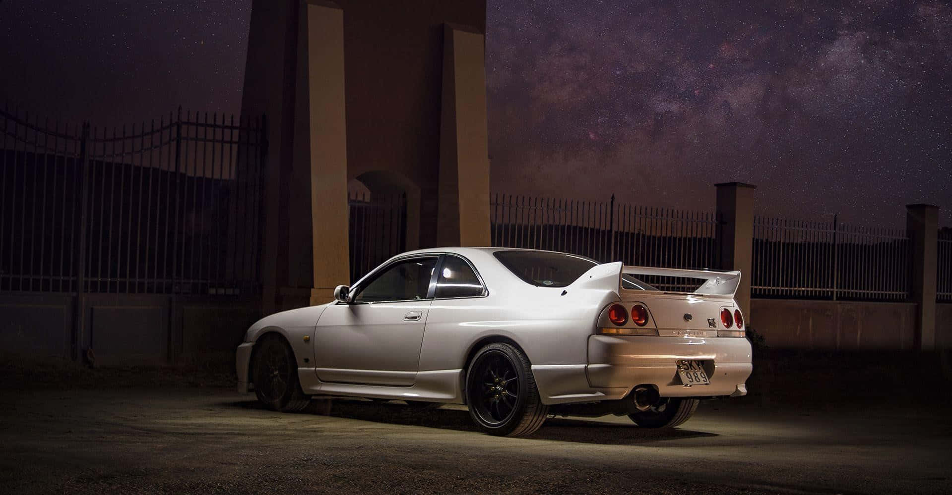R33 GT-R 桌布 壁紙 — 一輛銀色跑車在聚光燈下閃閃發光，背景是繁星點點的夜空，籬笆和建築物上方可見銀河。