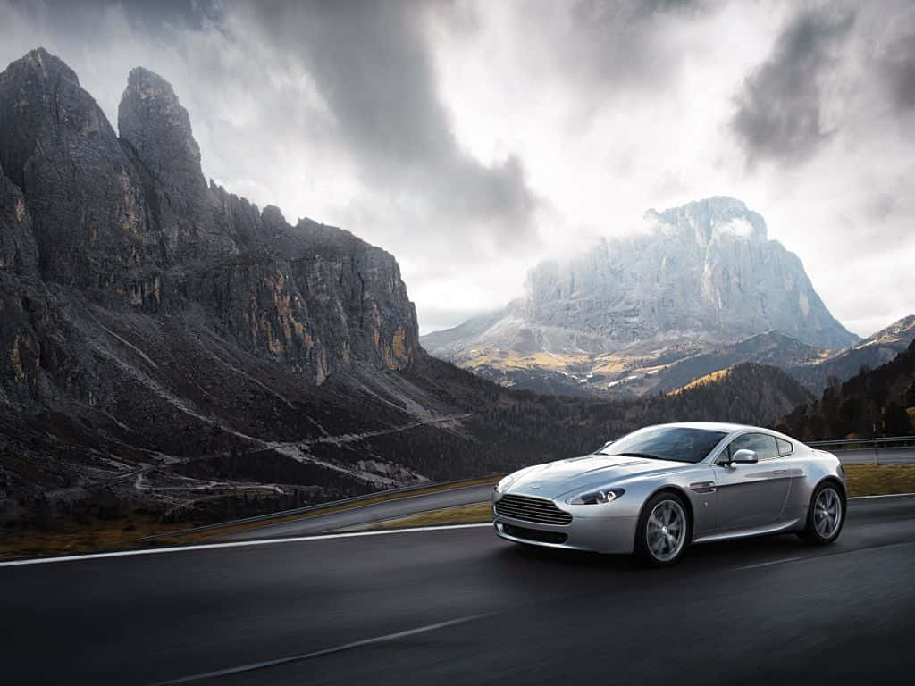 Aston Martin V12 Vantage 桌布 壁紙 — 一輛銀色跑車在蜿蜒的山路上行駛，背景是戲劇性的多雲天空和聳立的岩石山峰。