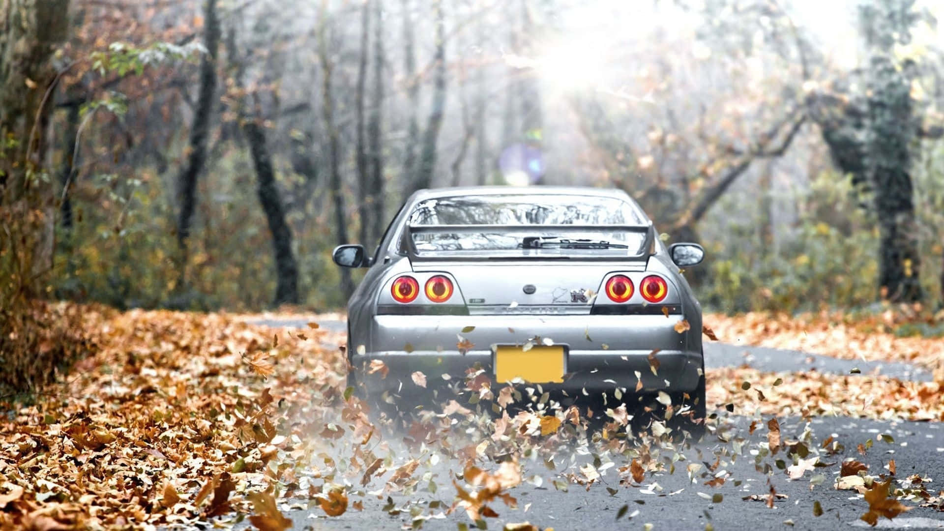 R33 GT-R 桌布 壁紙 — 一輛銀色跑車在霧氣瀰漫的森林道路上揚起秋葉，陽光透過光禿的樹木灑落。