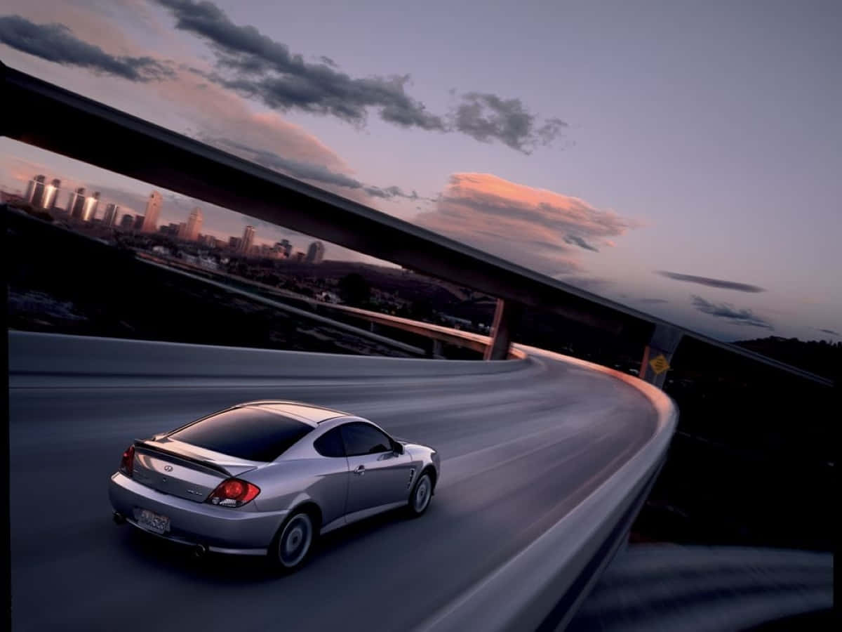 Hyundai Tiburon 桌布 壁紙 — 一輛銀色跑車在黃昏時分沿著蜿蜒的高速公路疾馳，遠處的城市天際線閃爍著光芒。