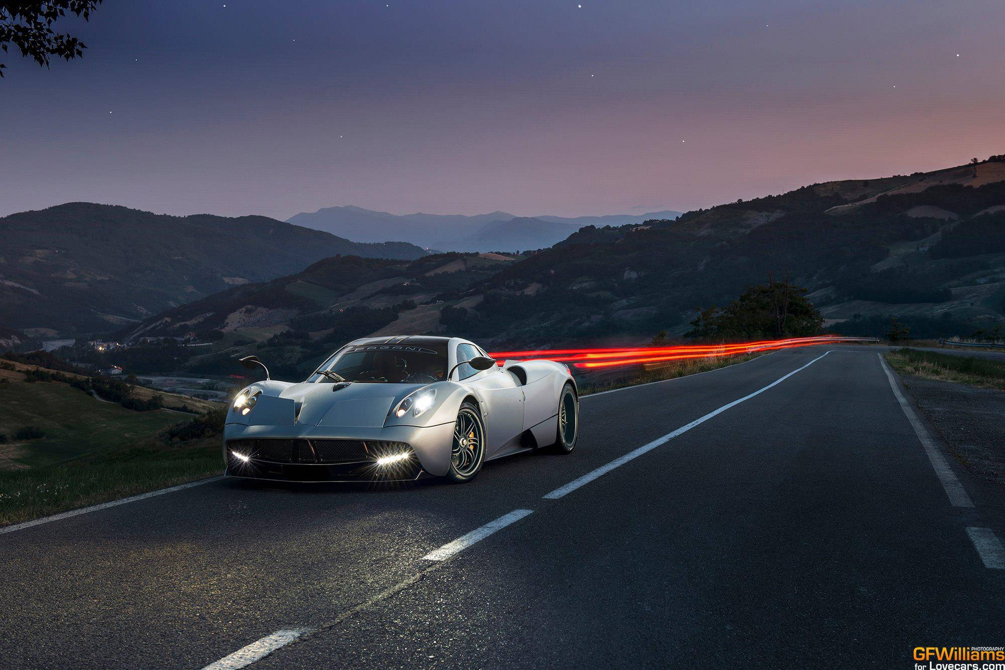 Pagani 桌布 壁紙 — 一輛銀色跑車在黃昏時分沿著蜿蜒的山路滑行，身後留下紅色光跡。