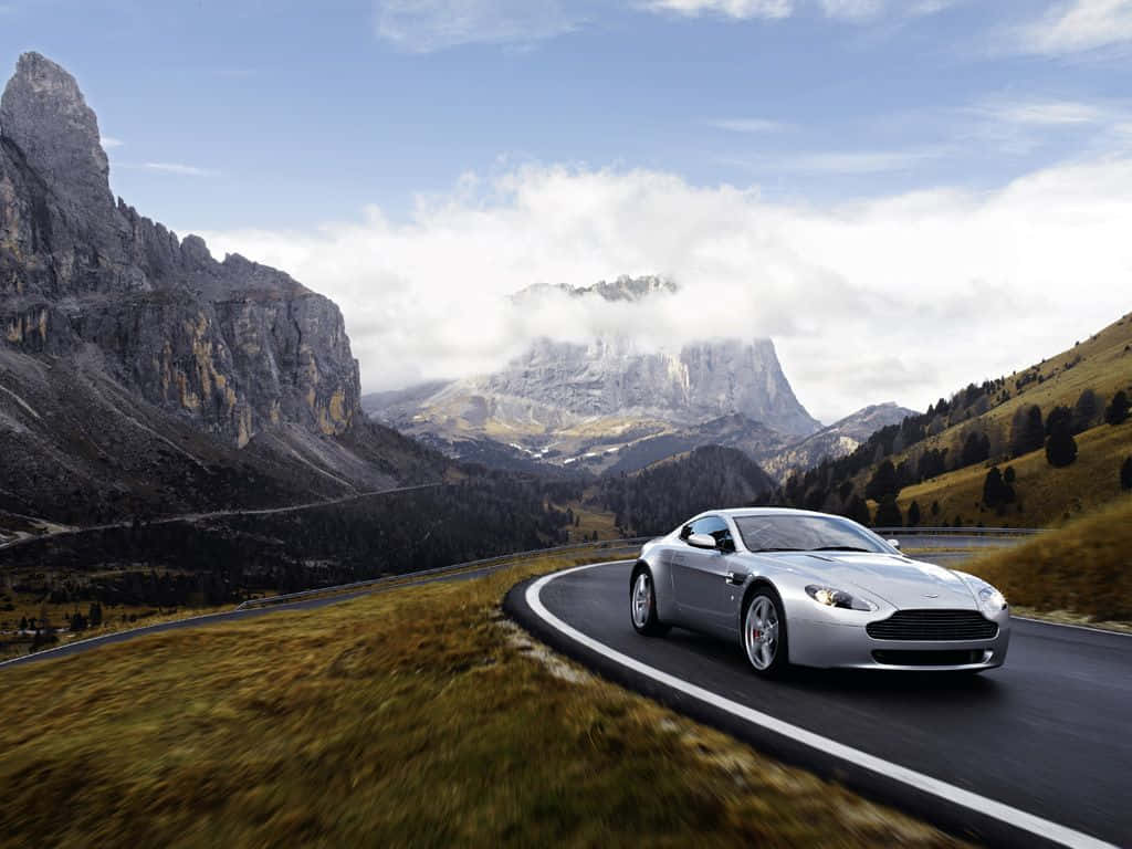 Aston Martin V12 Vantage 桌布 壁紙 — 一輛銀色跑車沿著蜿蜒的山路行駛，背景是戲劇性的山峰和雲彩。