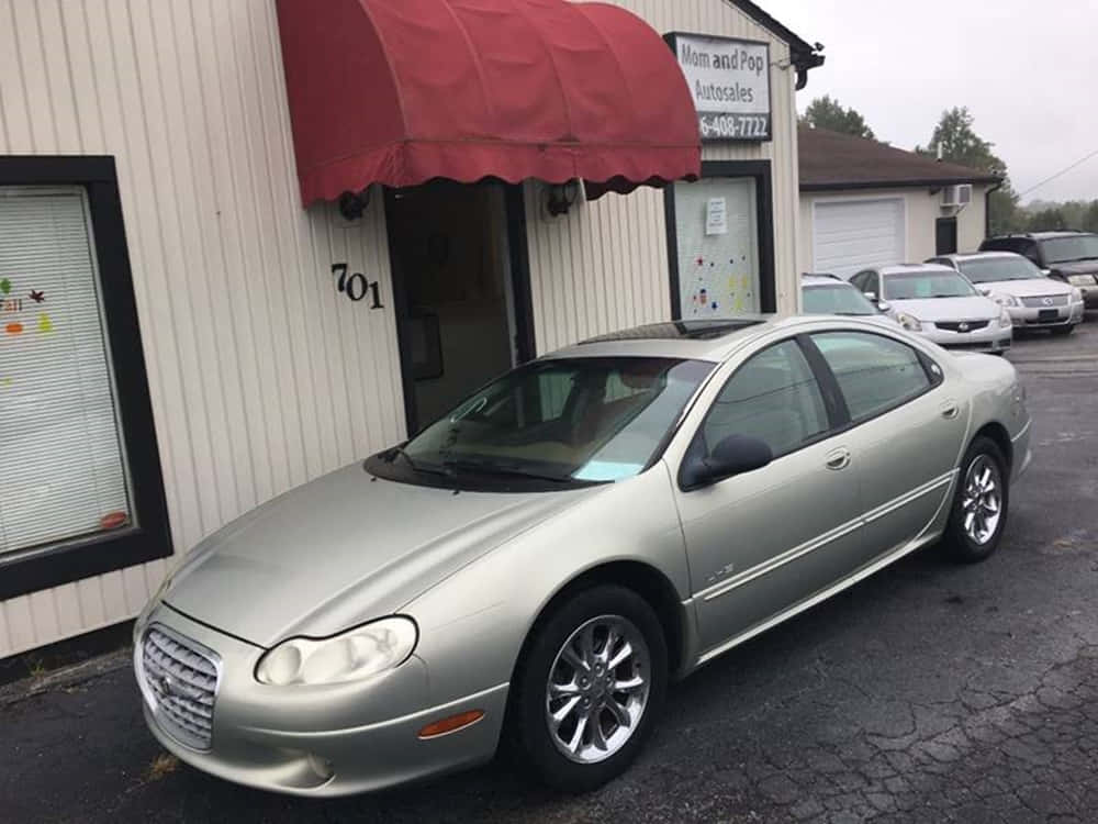 Chrysler Lhs 桌布 壁紙 — 一輛銀色轎車停在米色建築前，建築有紅色遮陽篷，天空多雲。