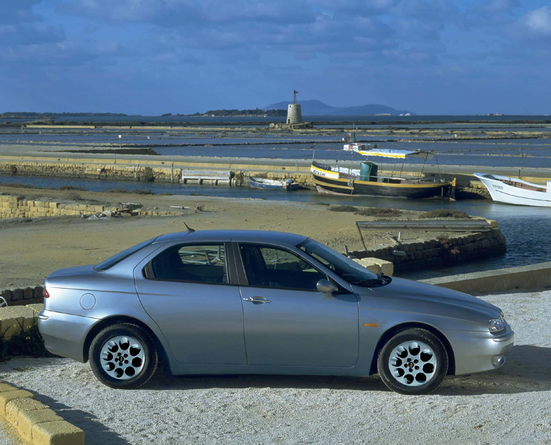 Alfa Romeo 156 桌布 壁紙 — 一輛銀色轎車停泊在碎石上，旁邊是港口，有船隻和遠處的燈塔，天空局部多雲。
