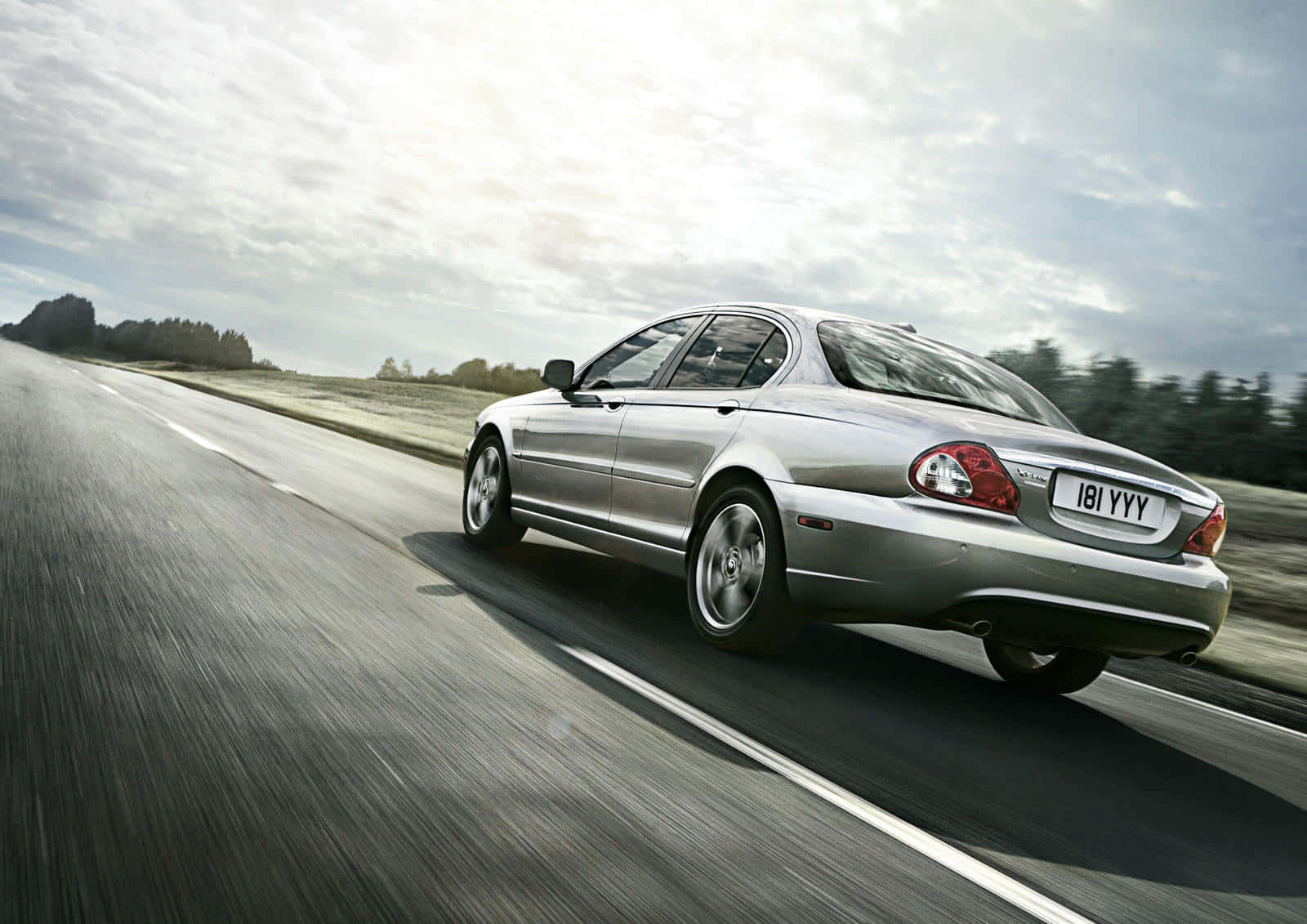 Jaguar X-type 桌布 壁紙 — 一輛銀色轎車在多雲的天空下於鄉村高速公路上疾馳，動態模糊強調了速度。