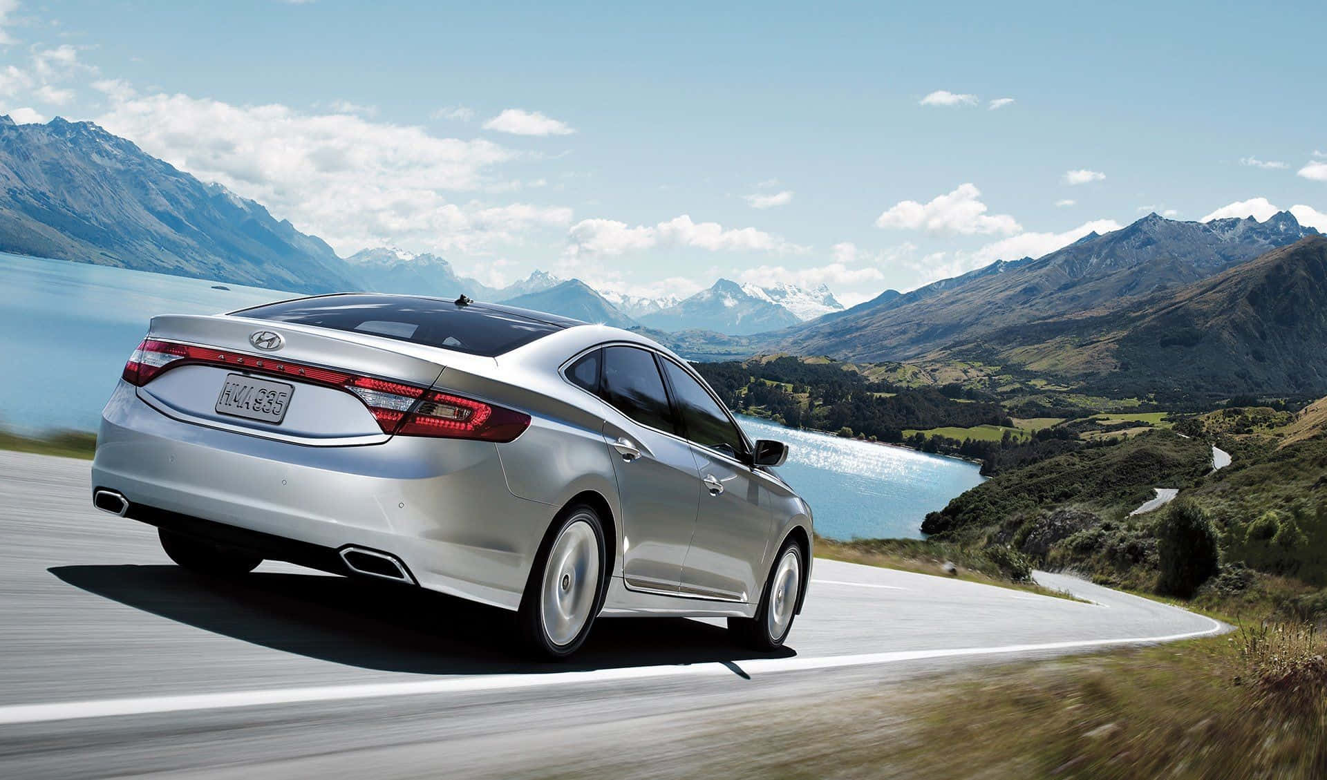 Hyundai Azera 桌布 壁紙 — 一輛銀色轎車在晴朗的天空下，沿著蜿蜒的山路行駛，旁邊是寧靜的藍色湖泊。