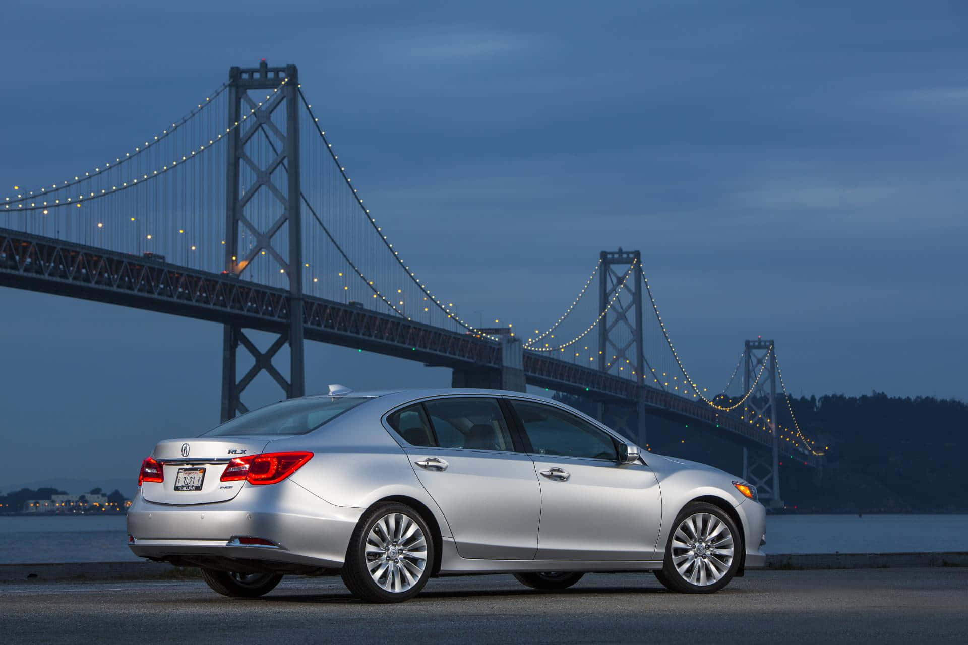 Acura Rlx 桌布 壁紙 — 一輛銀色轎車在黃昏時停泊，身後是燈火通明的海灣大橋，矗立於平靜的水面上。