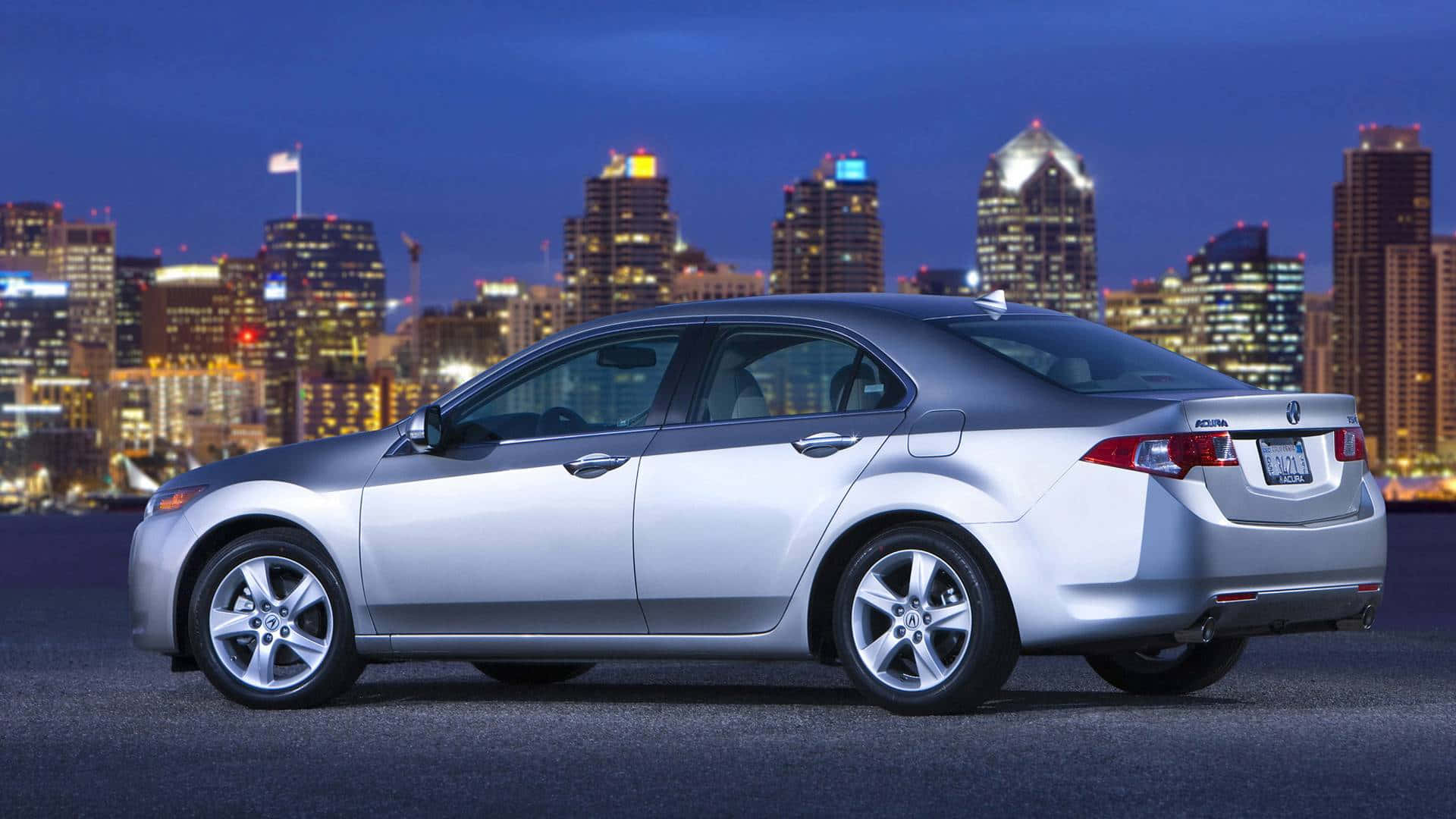 Acura Tsx 桌布 壁紙 — 一輛銀色轎車在黃昏時停泊在屋頂上，背後是充滿活力的城市天際線閃耀著光芒。