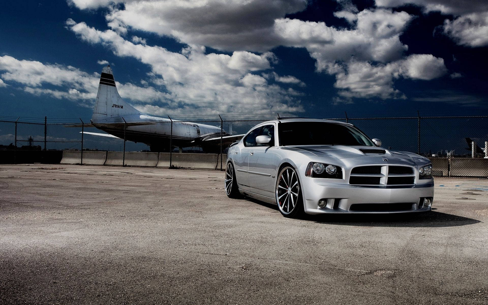 Dodge 桌布 壁紙 — 一輛銀色道奇Charger停在裂開的柏油路上，圍欄後面有一架大型軍用噴射機，天空戲劇性地多雲。
