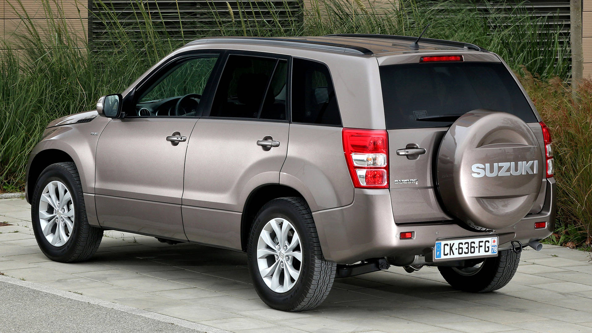 Suzuki 桌布 壁紙 — 一輛銀色鈴木 Grand Vitara SUV 停放在鋪砌路面上，後面有高高的草叢。