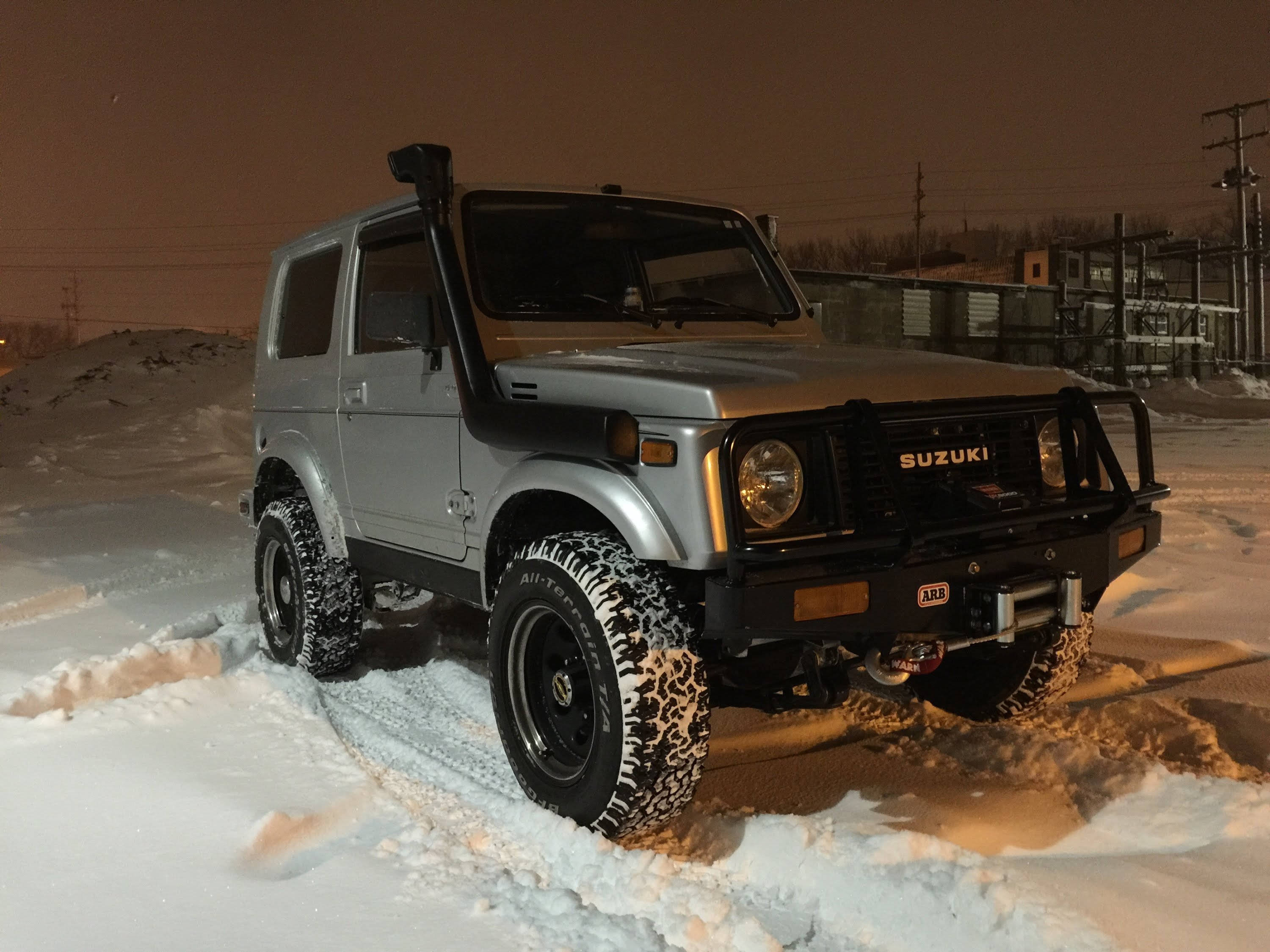 Suzuki 桌布 壁紙 — 一輛銀色鈴木Jimny SUV，輪胎被雪覆蓋，停在深雪中，夜空昏暗。