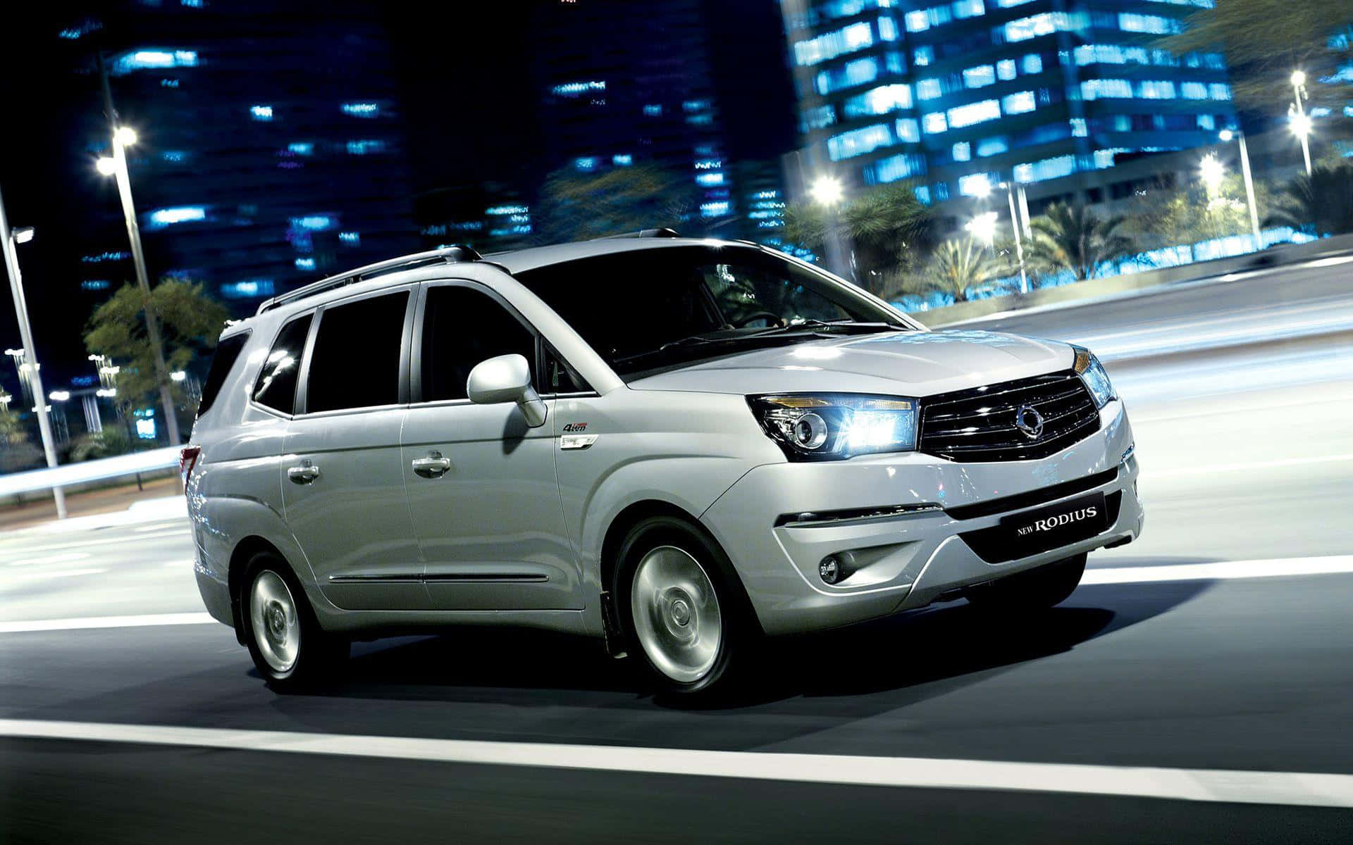 Ssangyong 桌布 壁紙 — 一輛銀色雙龍Rodius廂型車在霓虹燈閃爍的城市夜間高速行駛，留下模糊的動態軌跡。